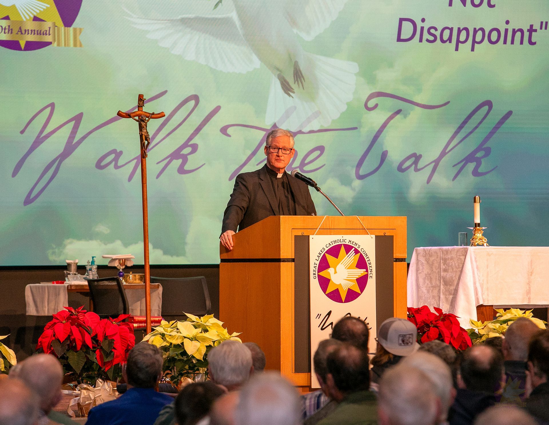 Fr. Rocky at the podium on stage with floral arrangements and the audience, under a “Walk the Talk” banner.