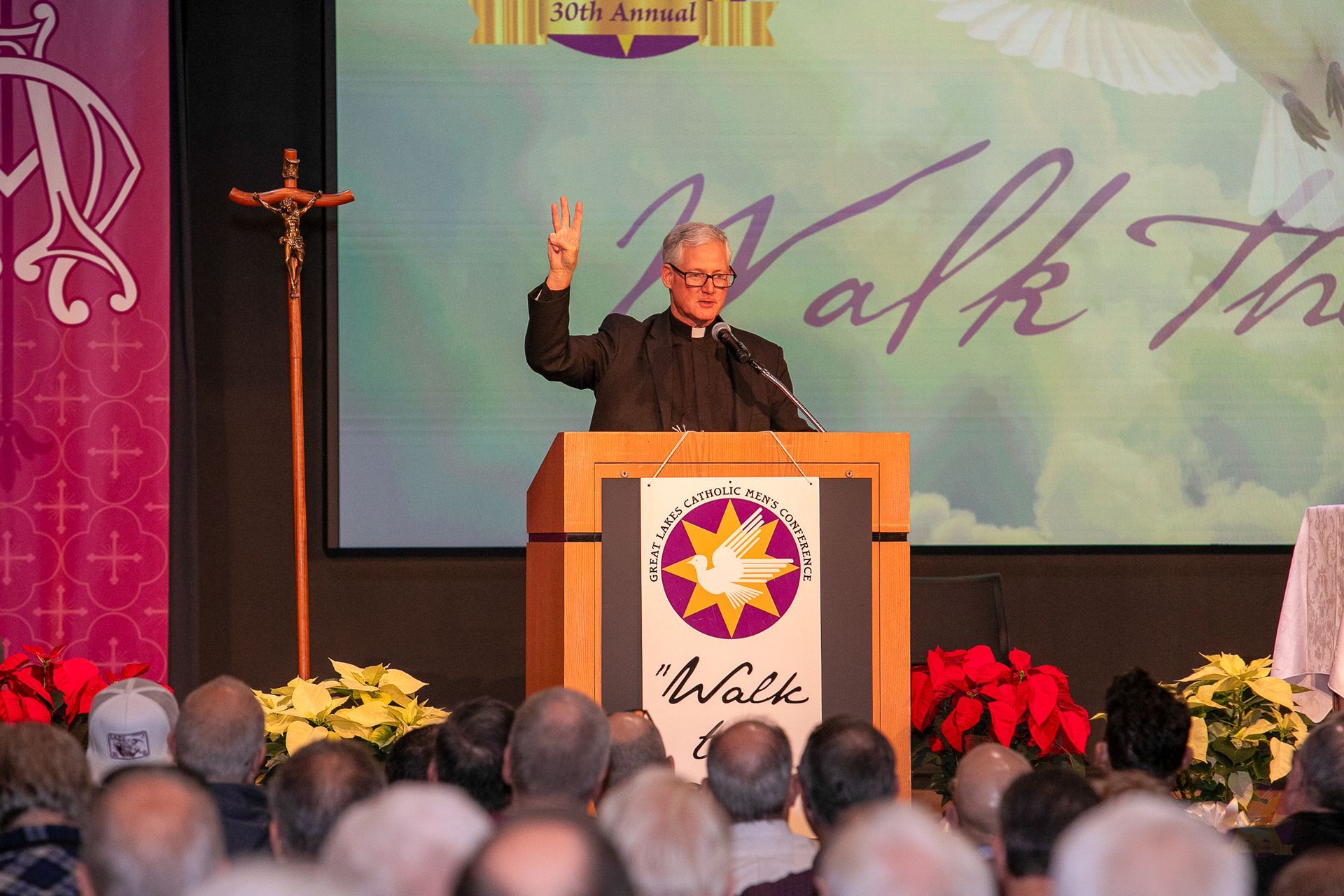 Fr. Rocky at podium with raised hand on stage, with audience and holiday decorations in front
