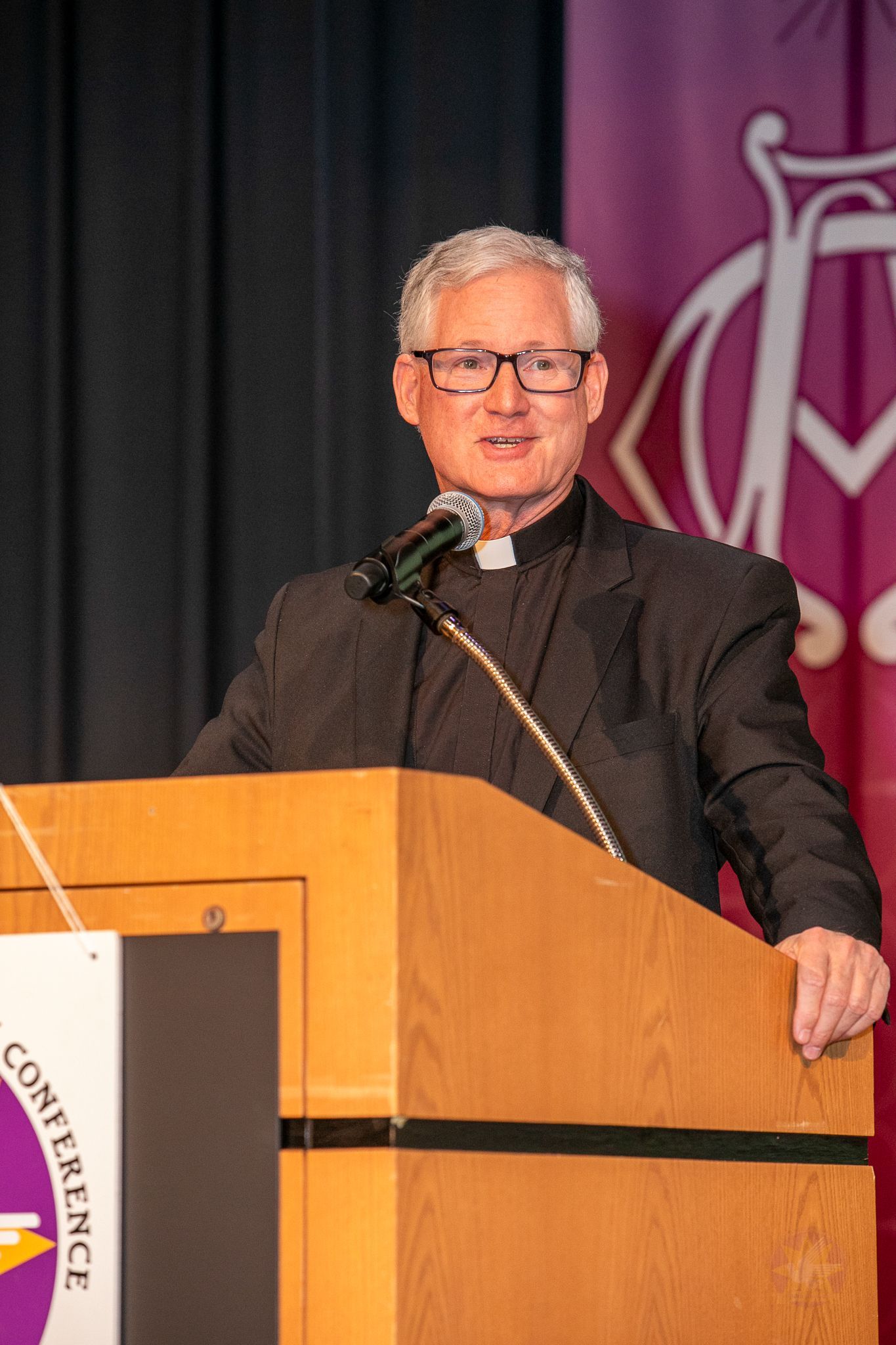 Fr. Rocky speaking at a podium with microphone, in front of a purple backdrop with a logo
