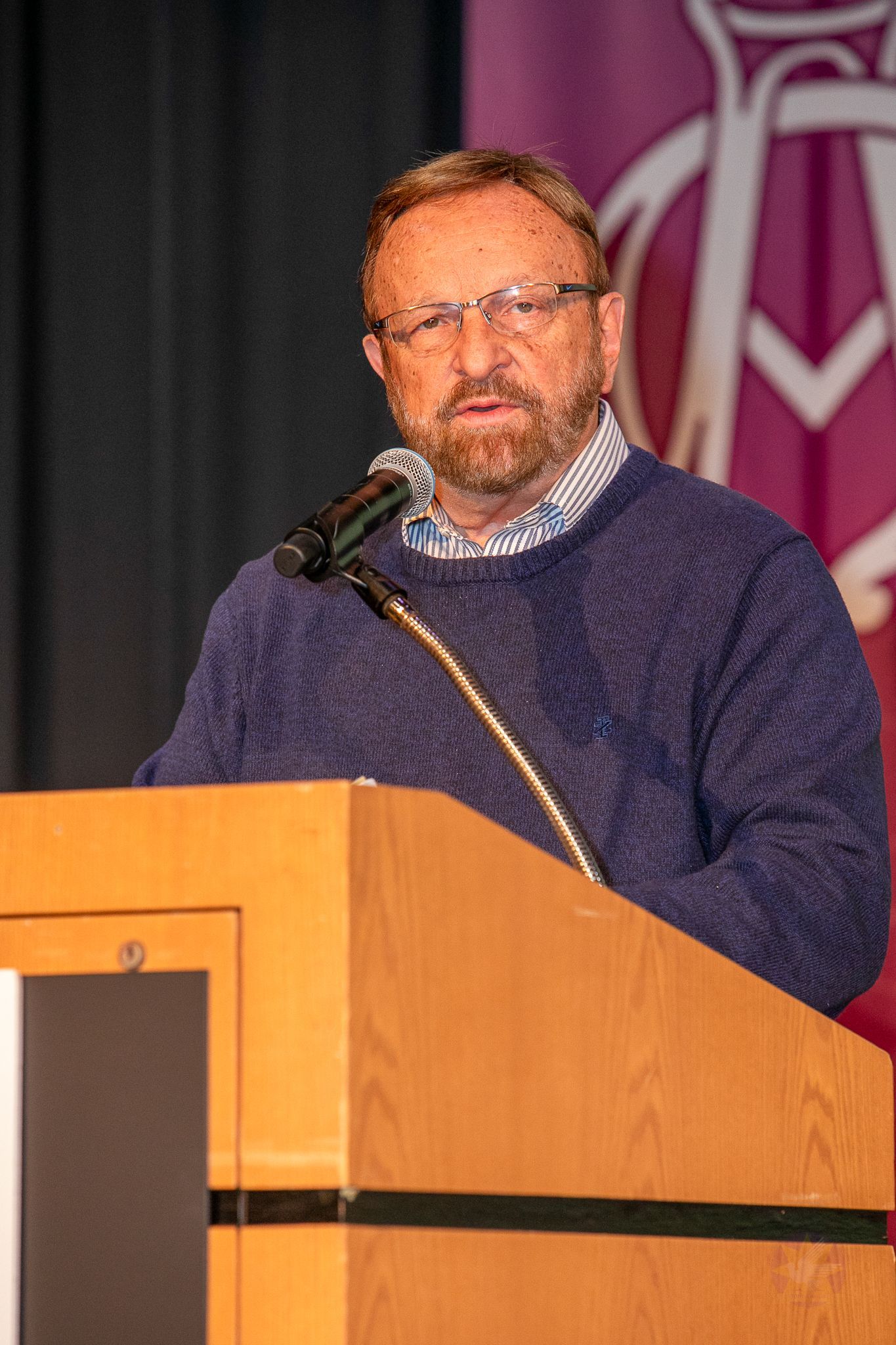 Wayne Messmer speaking at a podium with a microphone, wearing a navy sweater and glasses, on a stage with a maroon backdrop.