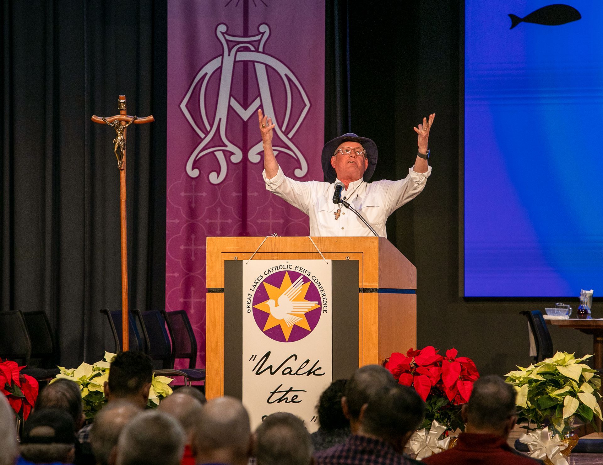 Steve Ray speaking at a podium on stage, arms raised, with a purple banner and audience in front