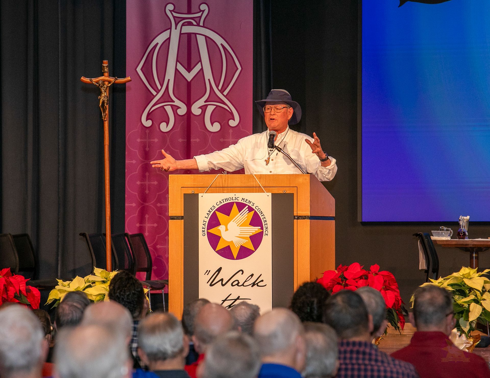Steve Ray at podium on stage, gesturing before a seated audience, with purple and blue backdrop and floral arrangements