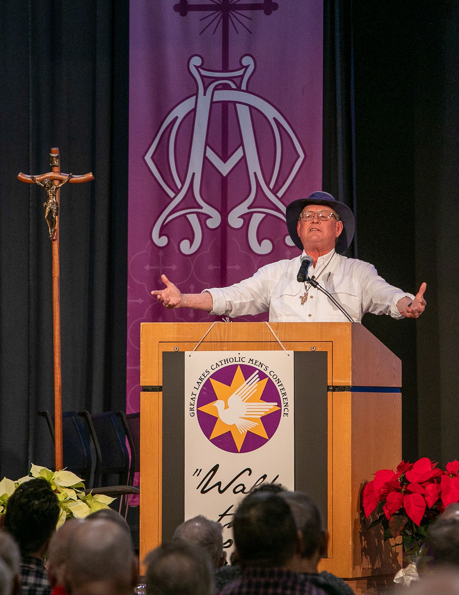 Speaker at podium on stage with arms outstretched, purple curtain backdrop and church emblem banner.