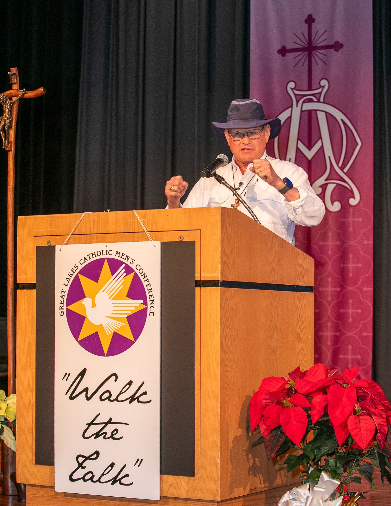 Steve Ray  speaking at a podium on stage with a “Walk the Walk” sign and red flowers