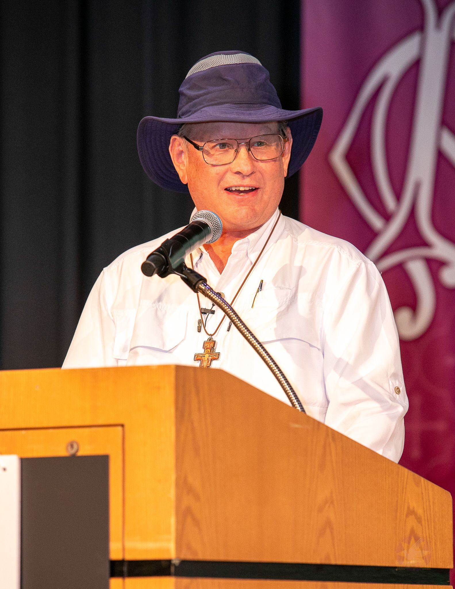 Steve Ray, in a hat, speaking at a podium with a microphone on a stage.
