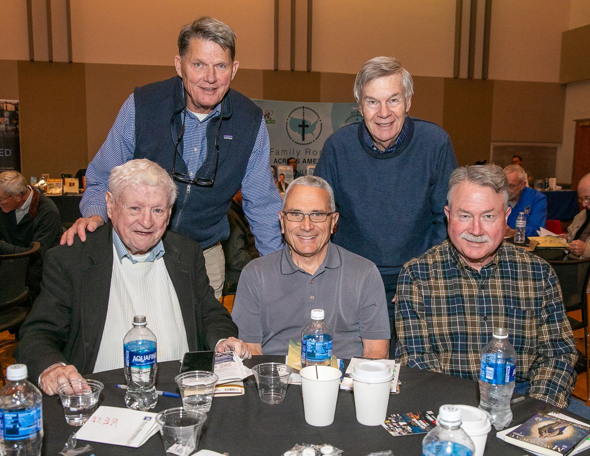 Five smiling men pose at a banquet table with drinks and water bottles in a conference hall.