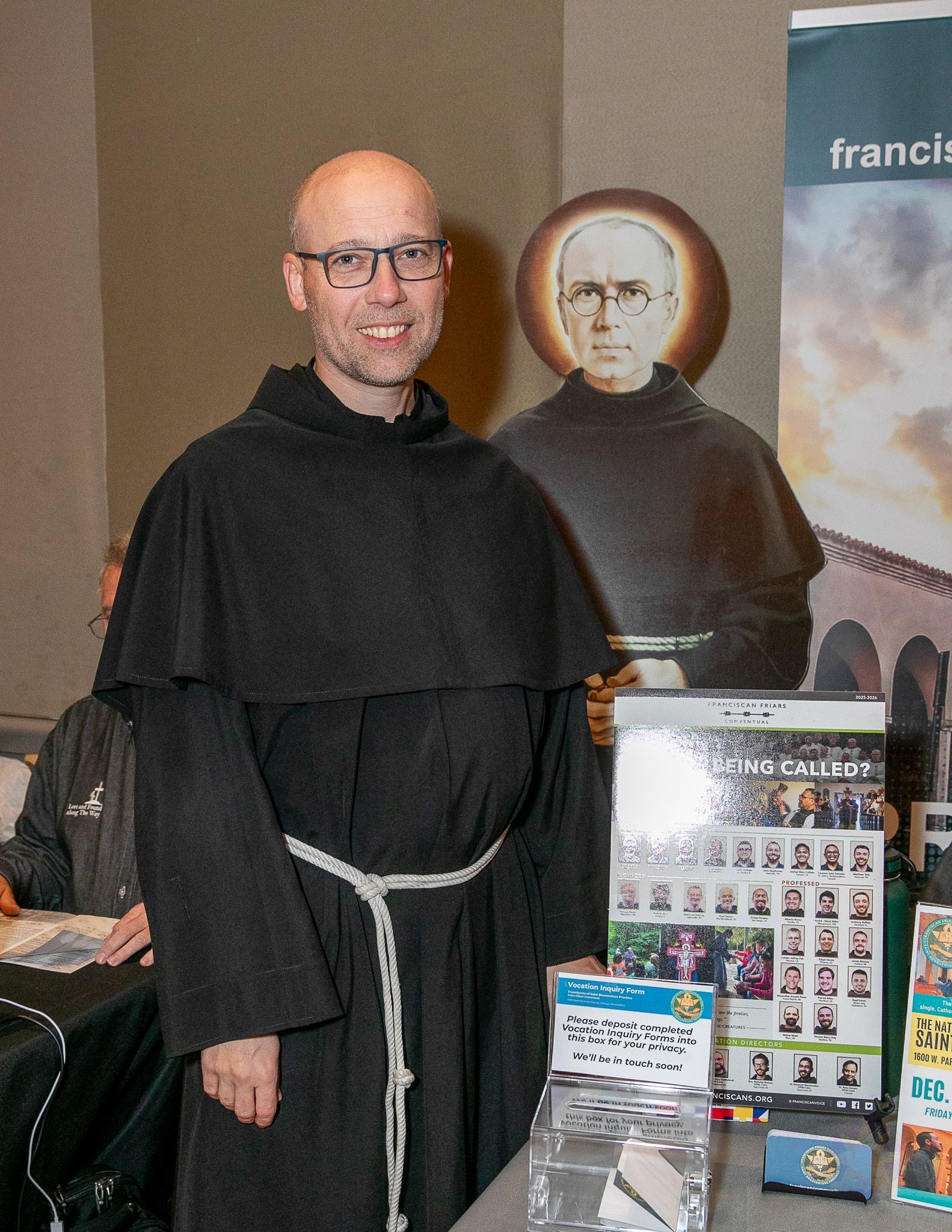 A Franciscan Brother stands beside a display table with a portrait and brochures.