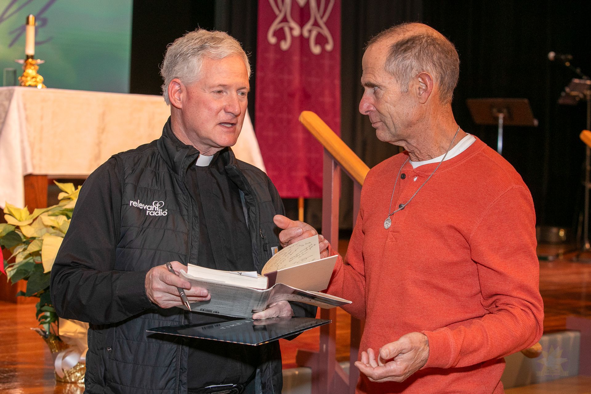 Two men speaking at a church altar, one holding a book and the other a paper, with candles and flowers in the background.