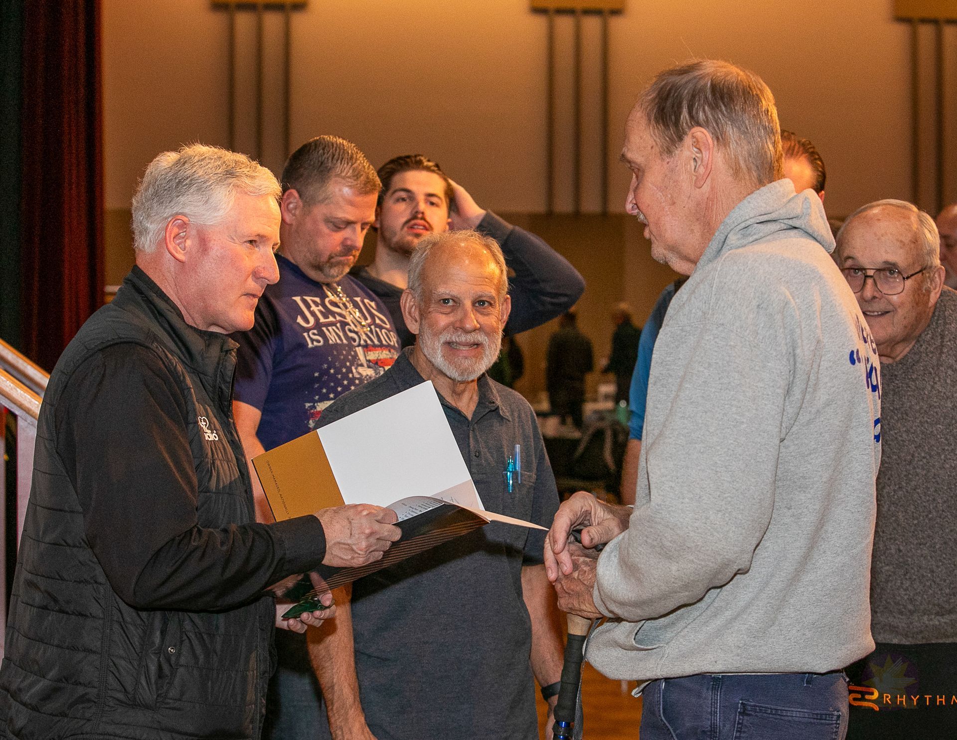 Two men shake hands at a Catholic men's conference while others stand nearby, one holding an open folder.