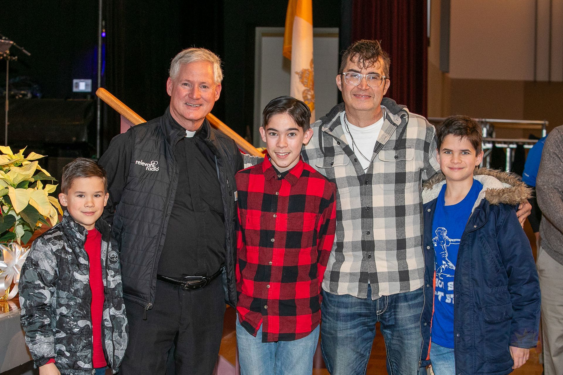Five people stand indoors, smiling, with a boy in a red plaid shirt at center.