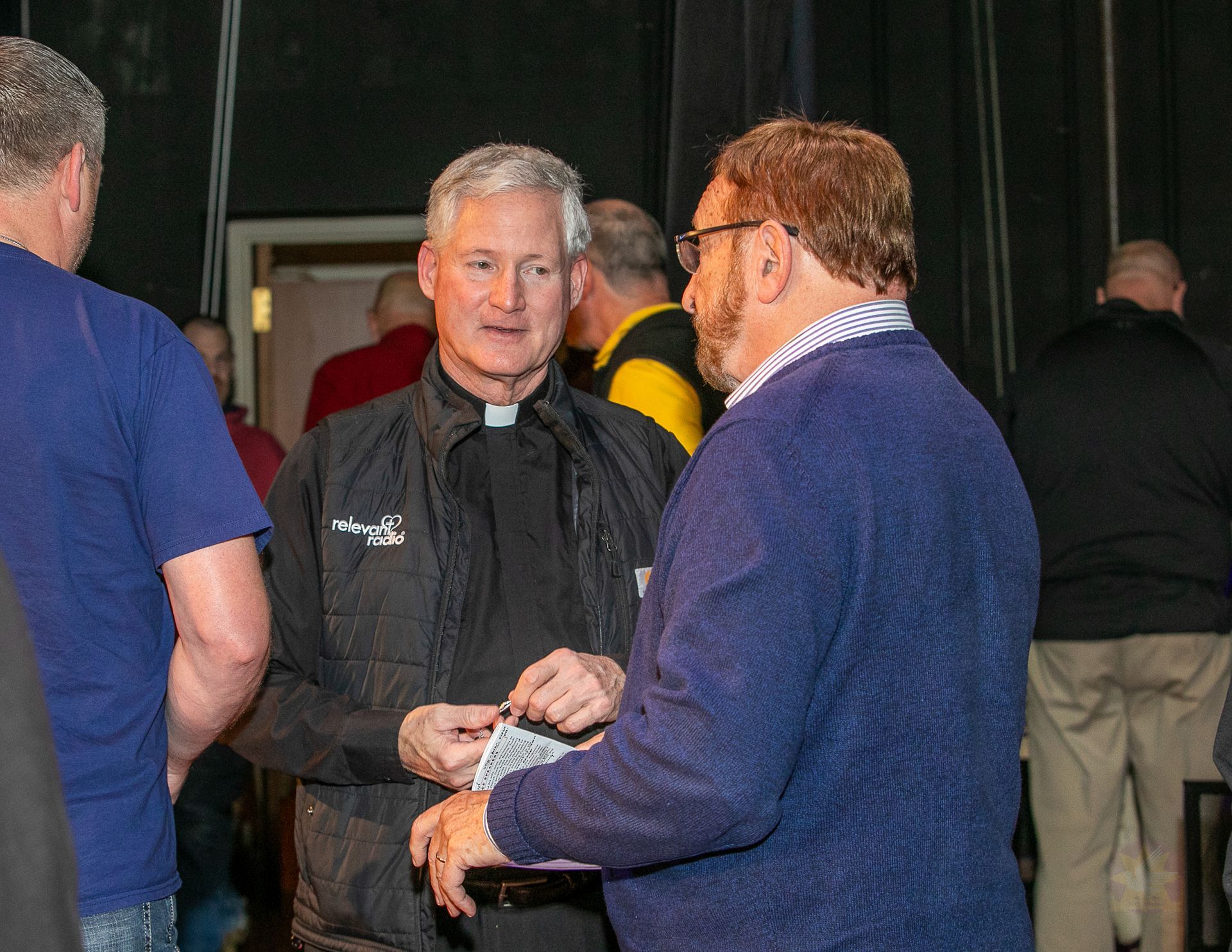 Three people conversing indoors, with a man in a black jacket facing the camera.