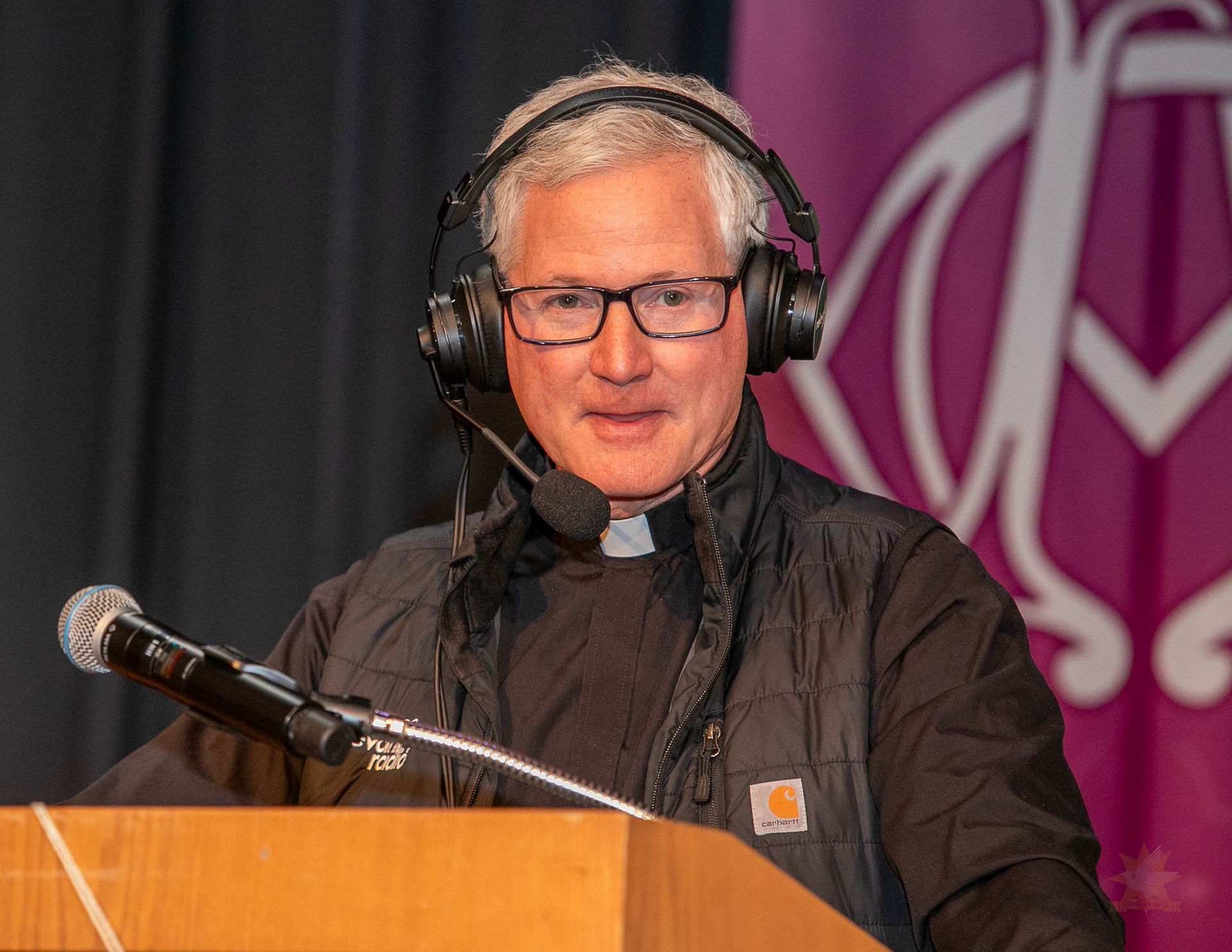 Fr. Rocky wearing headphones speaking at a podium with a microphone on stage