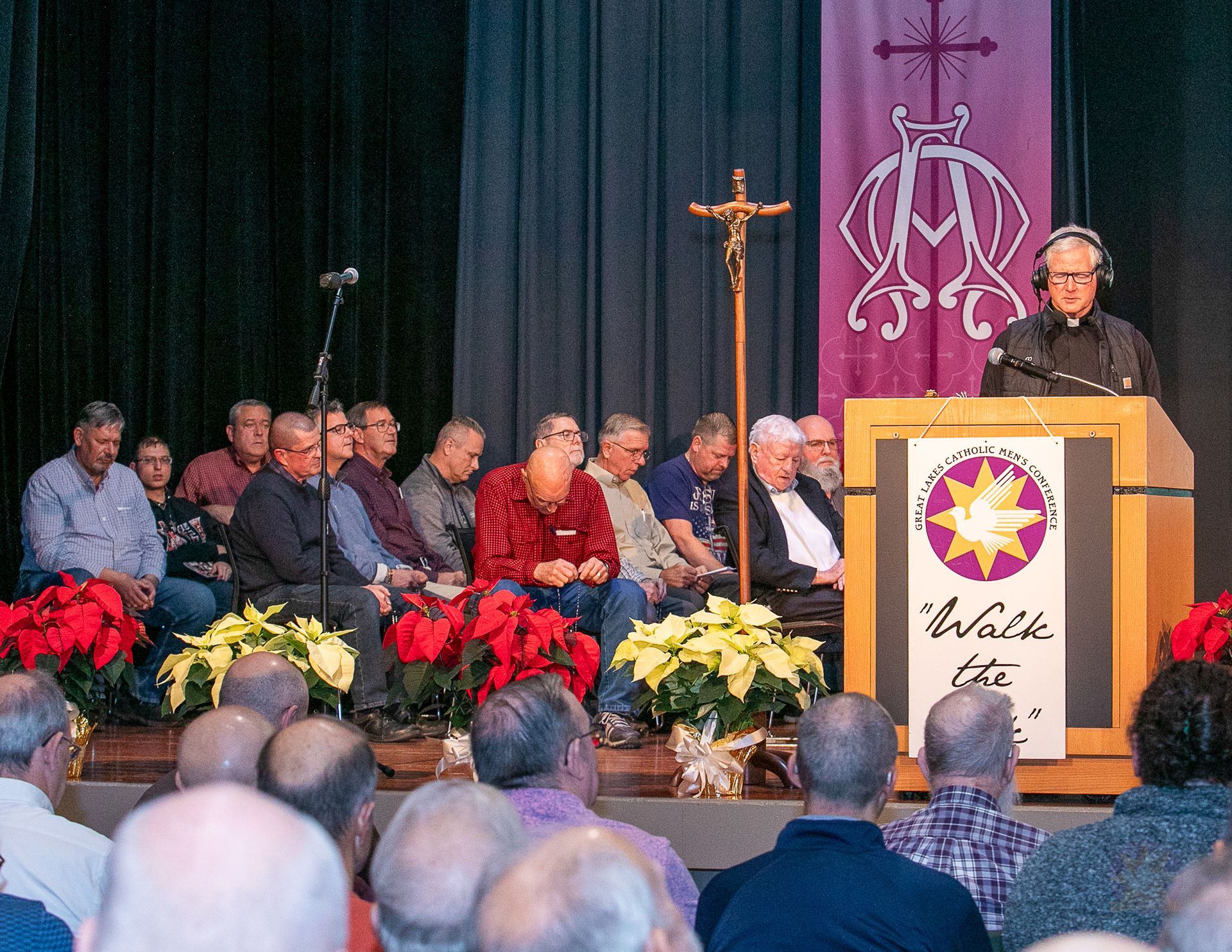 Fr. Rocky at podium addressing seated audience on stage, with flowers and a cross backdrop.