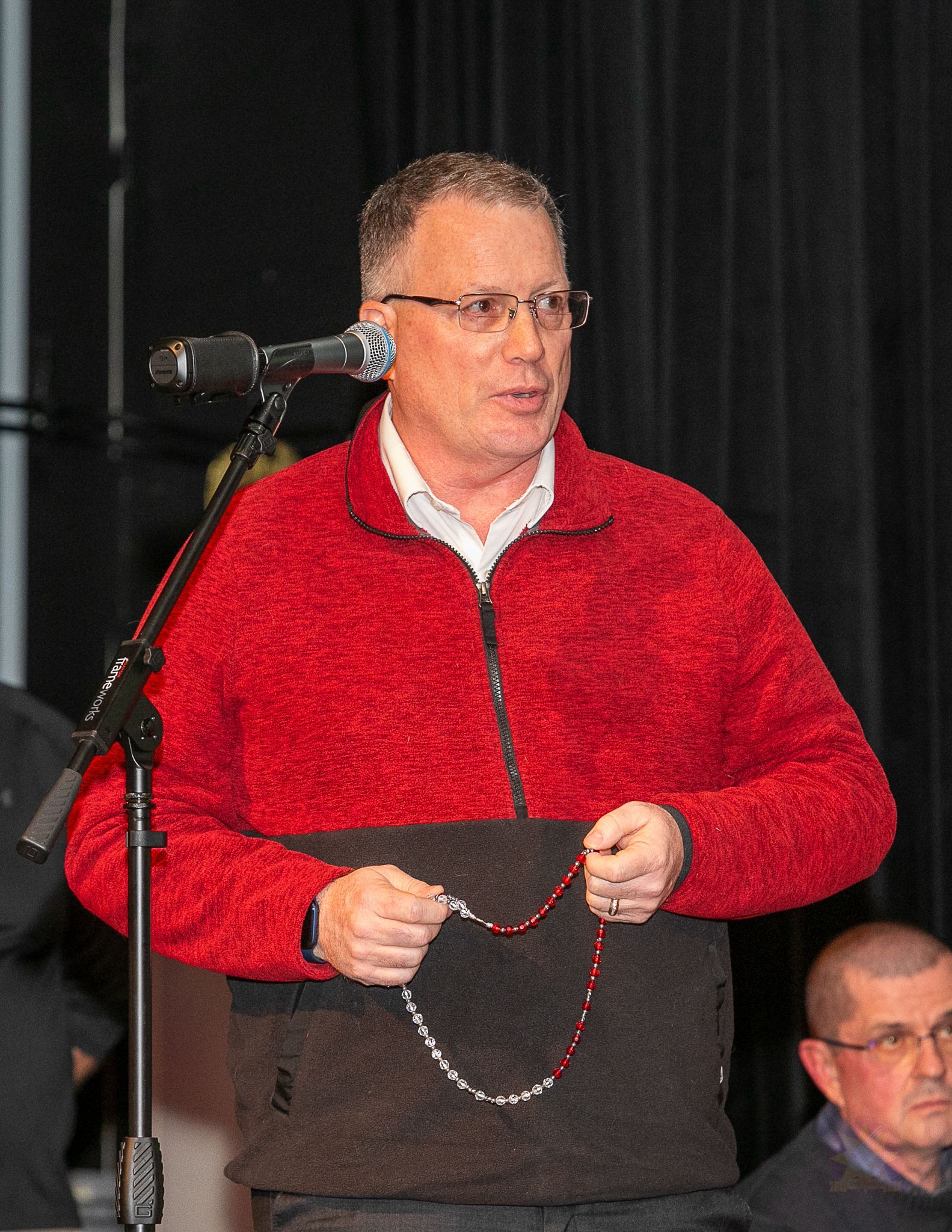 Man in red sweater speaking at a microphone, holding a rosary on a stage.