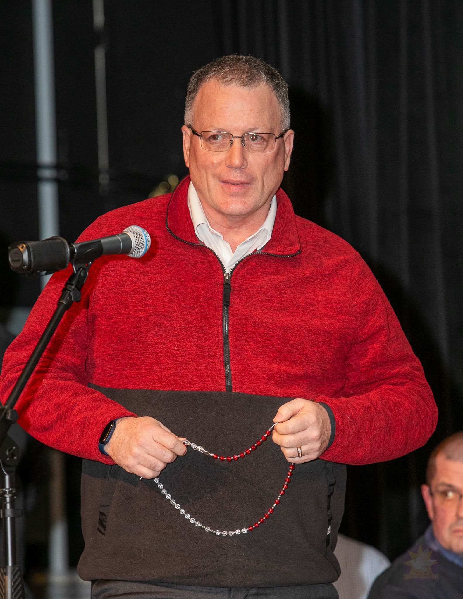 Man in a red sweater holding a beaded necklace beside microphones on a stage