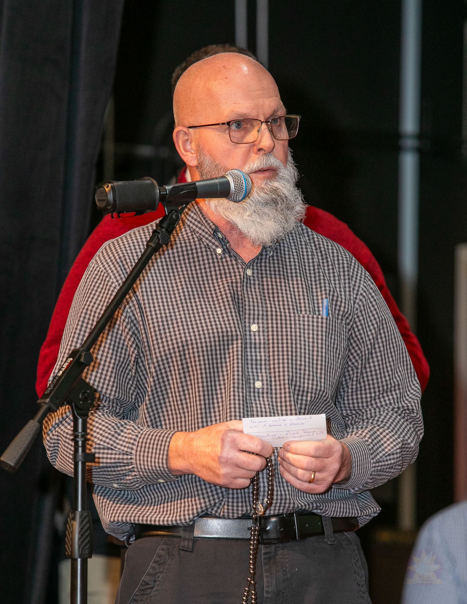 Bald man with gray beard speaking at a microphone, holding notes on stage, wearing a striped shirt and red jacket
