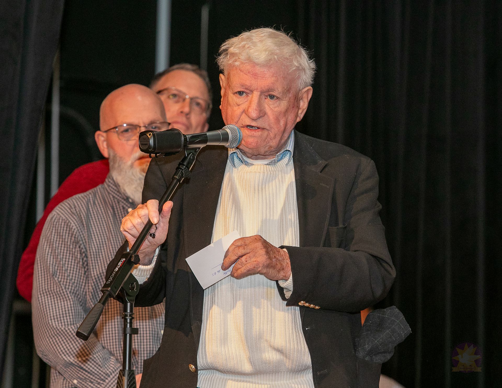Man speaking at a microphone on a stage, holding papers, with two people standing behind him.