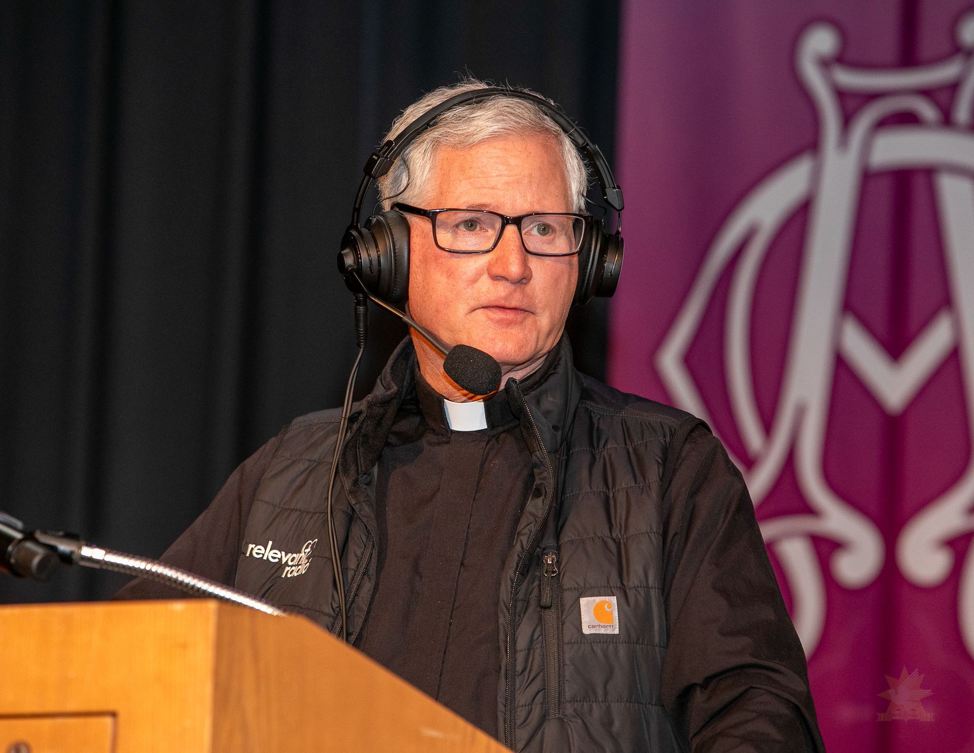 Fr. Rocky with headset microphone speaking at a podium on a stage with purple banner backdrop