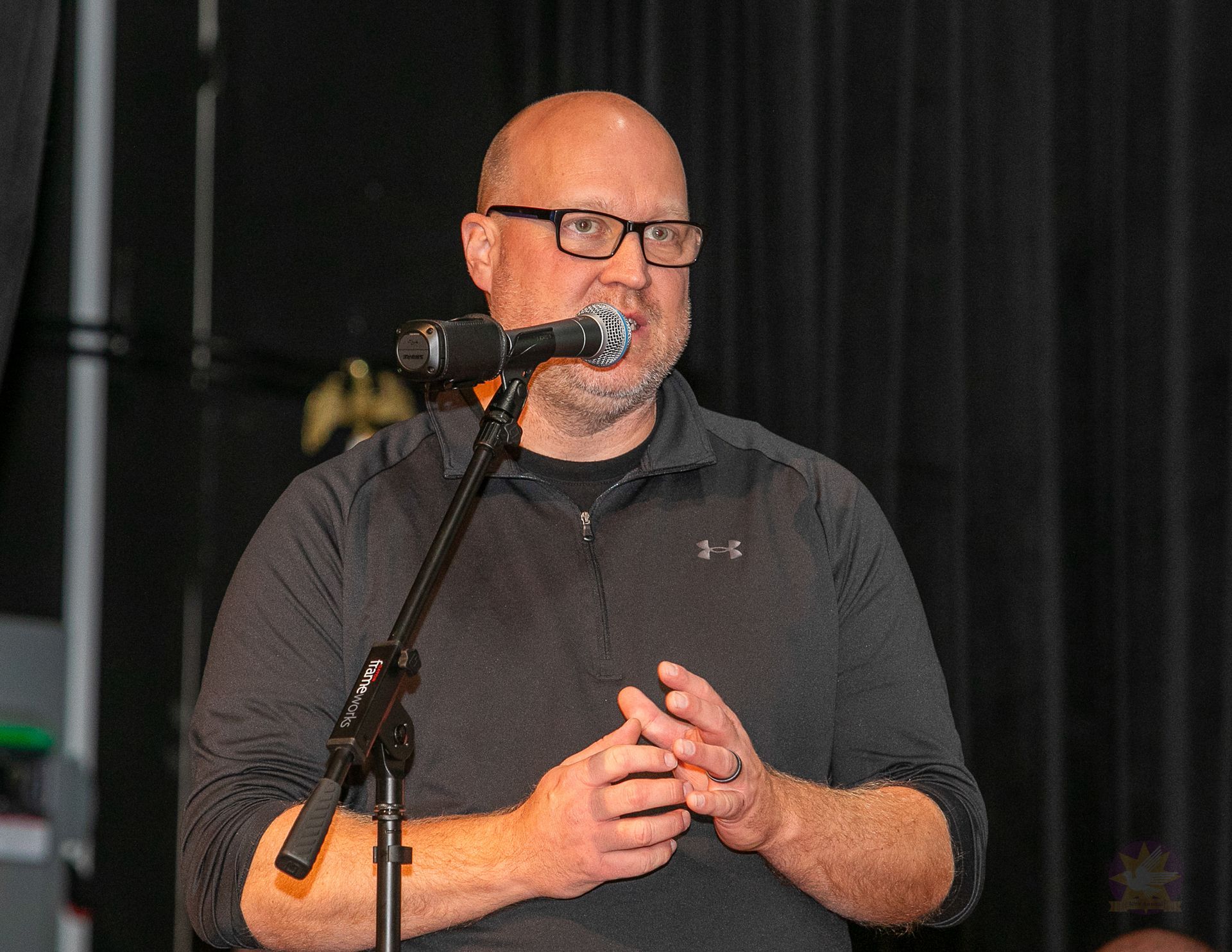Man speaking into a microphone on a stage, wearing a dark shirt against a black curtain backdrop.