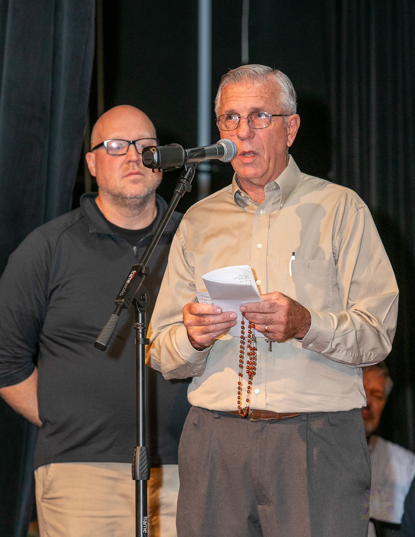 Man speaking into a microphone while reading from notes, with another man standing beside him on stage