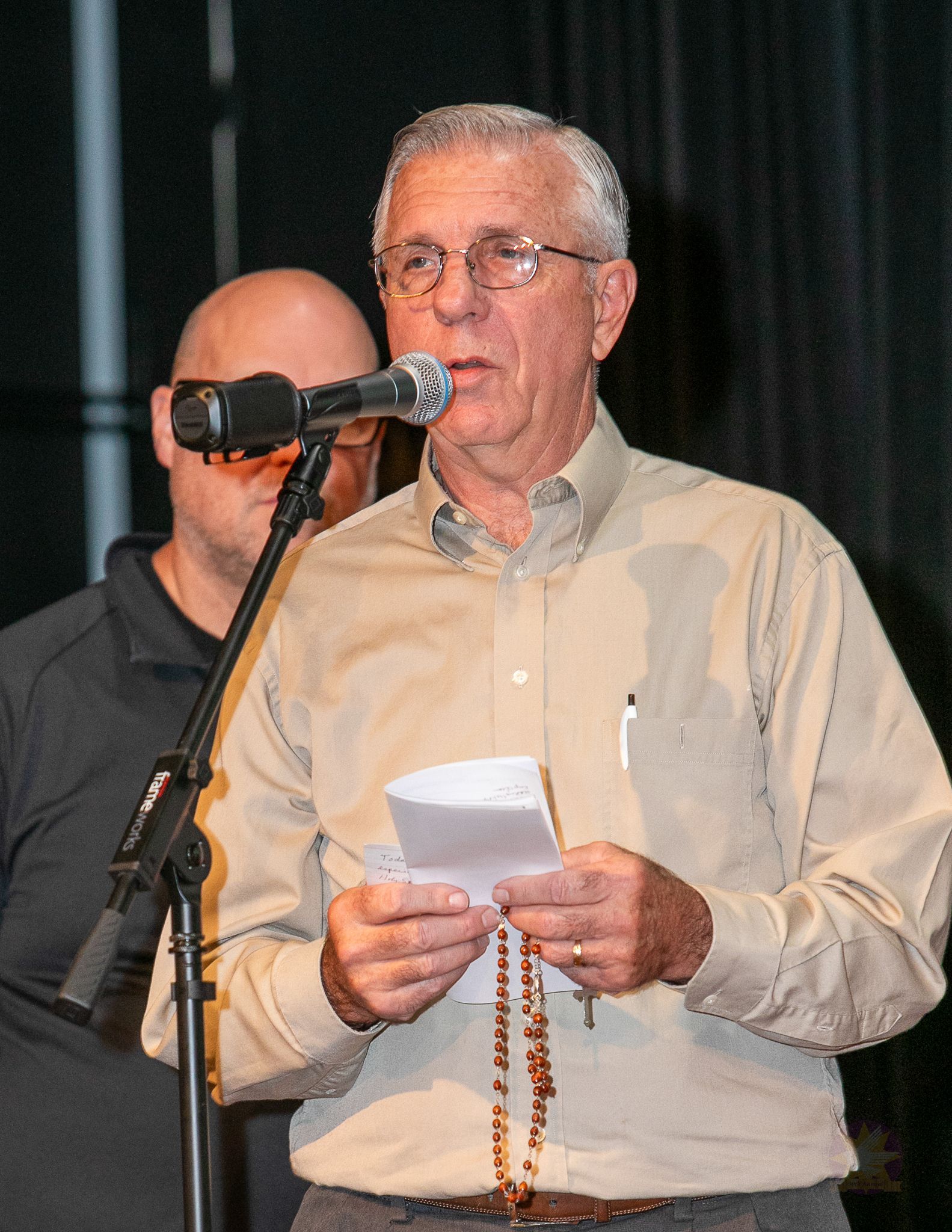 Man speaking at a microphone, holding notes and prayer beads, with another person partially visible behind him.