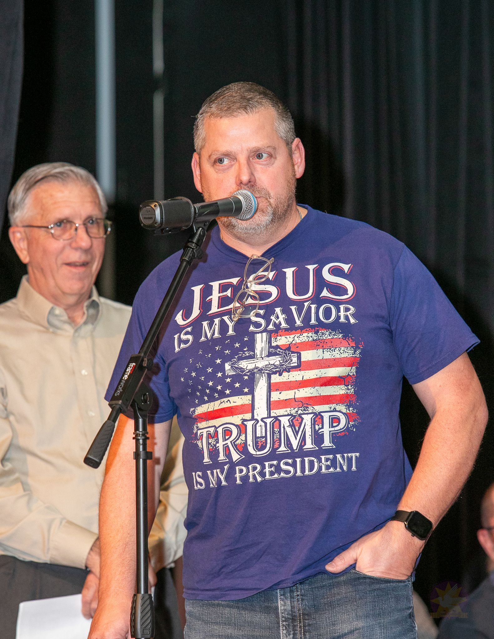 Man speaking at microphone in purple “Jesus is my Savior, Trump is my President” T-shirt, man behind him