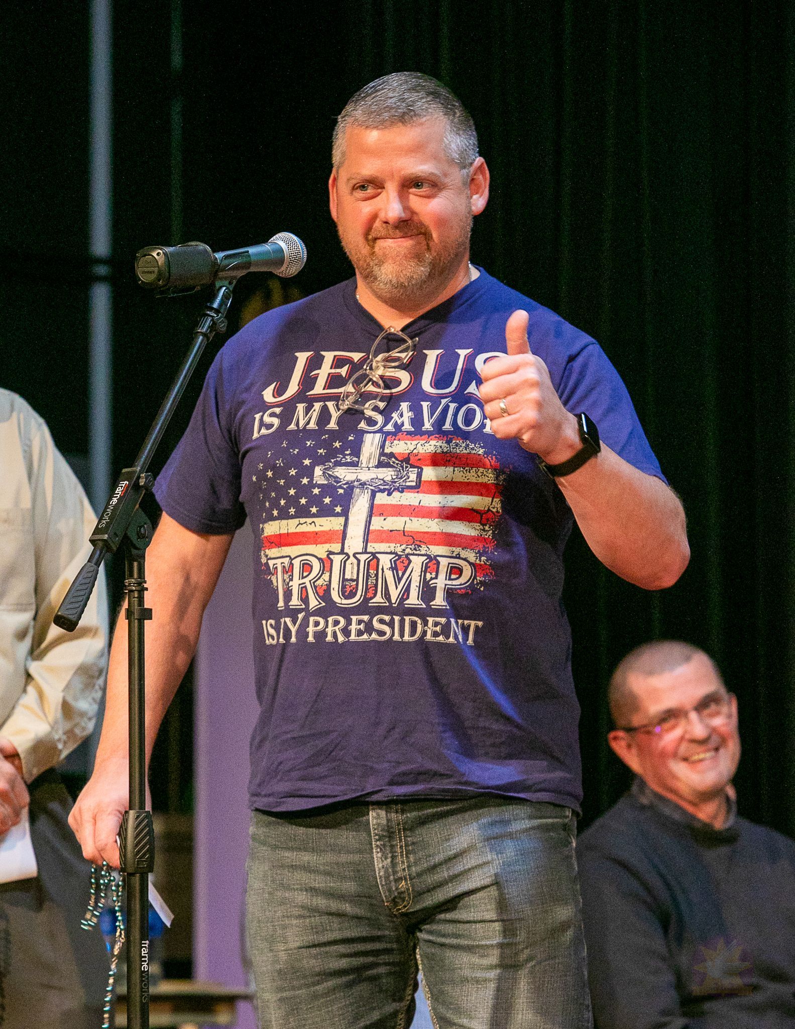 Man on stage giving a thumbs-up, wearing a dark graphic T-shirt, with a microphone and another man seated behind him.