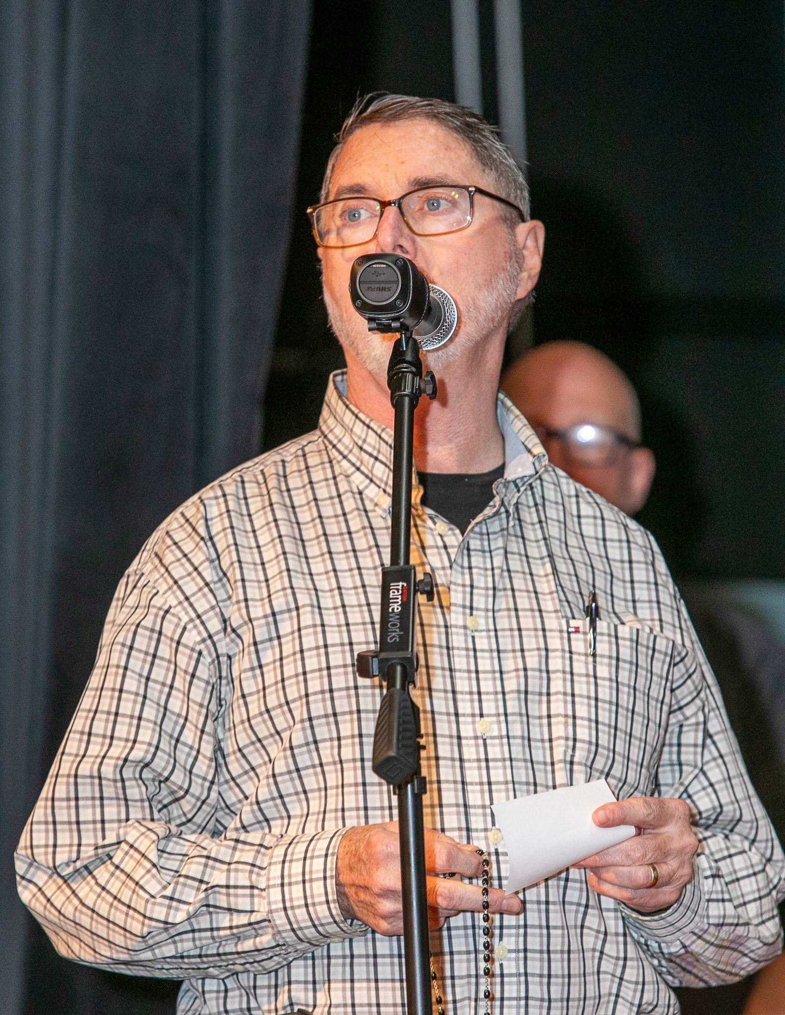 Man speaking into a microphone at a podium, holding papers on a stage with dark curtains.