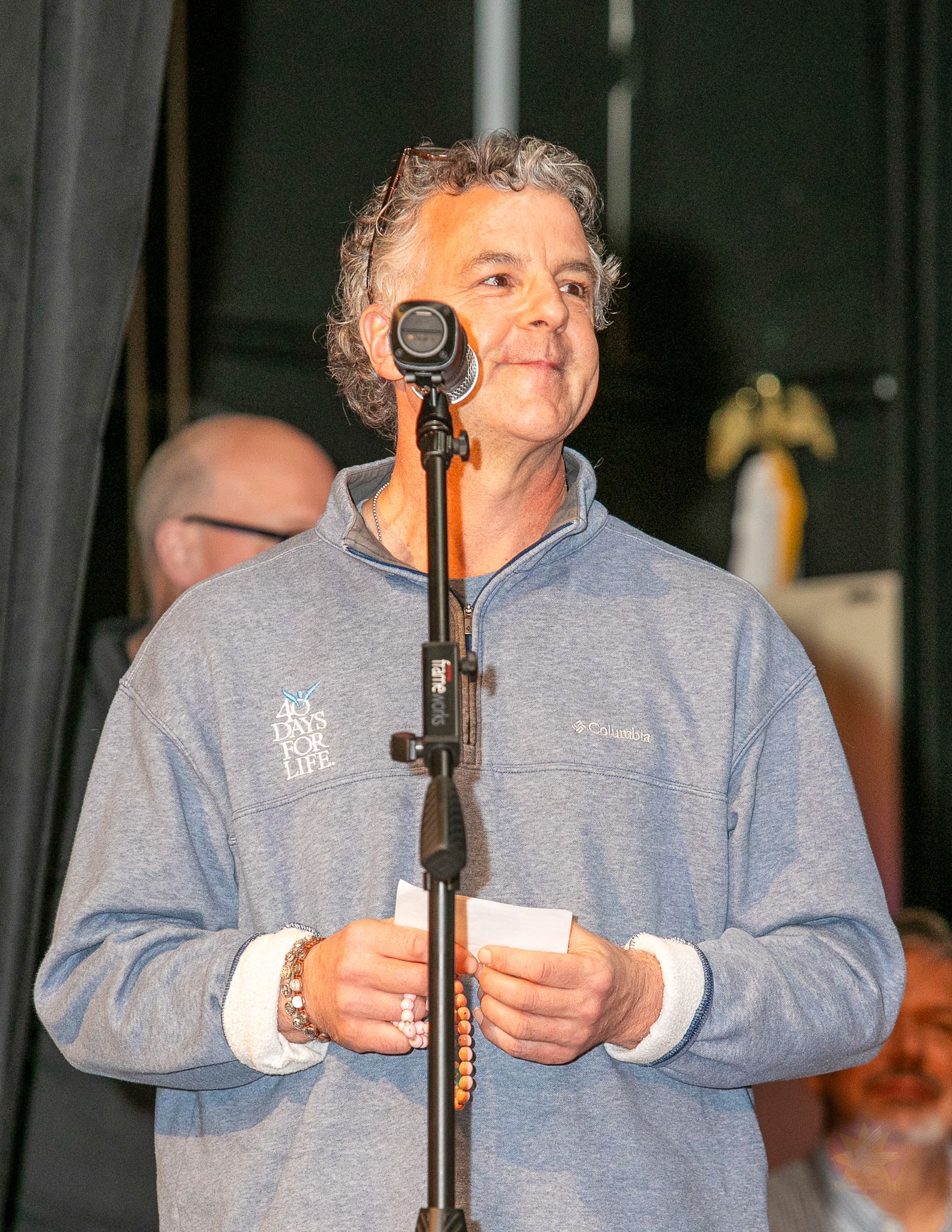 Man speaking at a microphone, holding note cards, wearing a blue shirt on stage