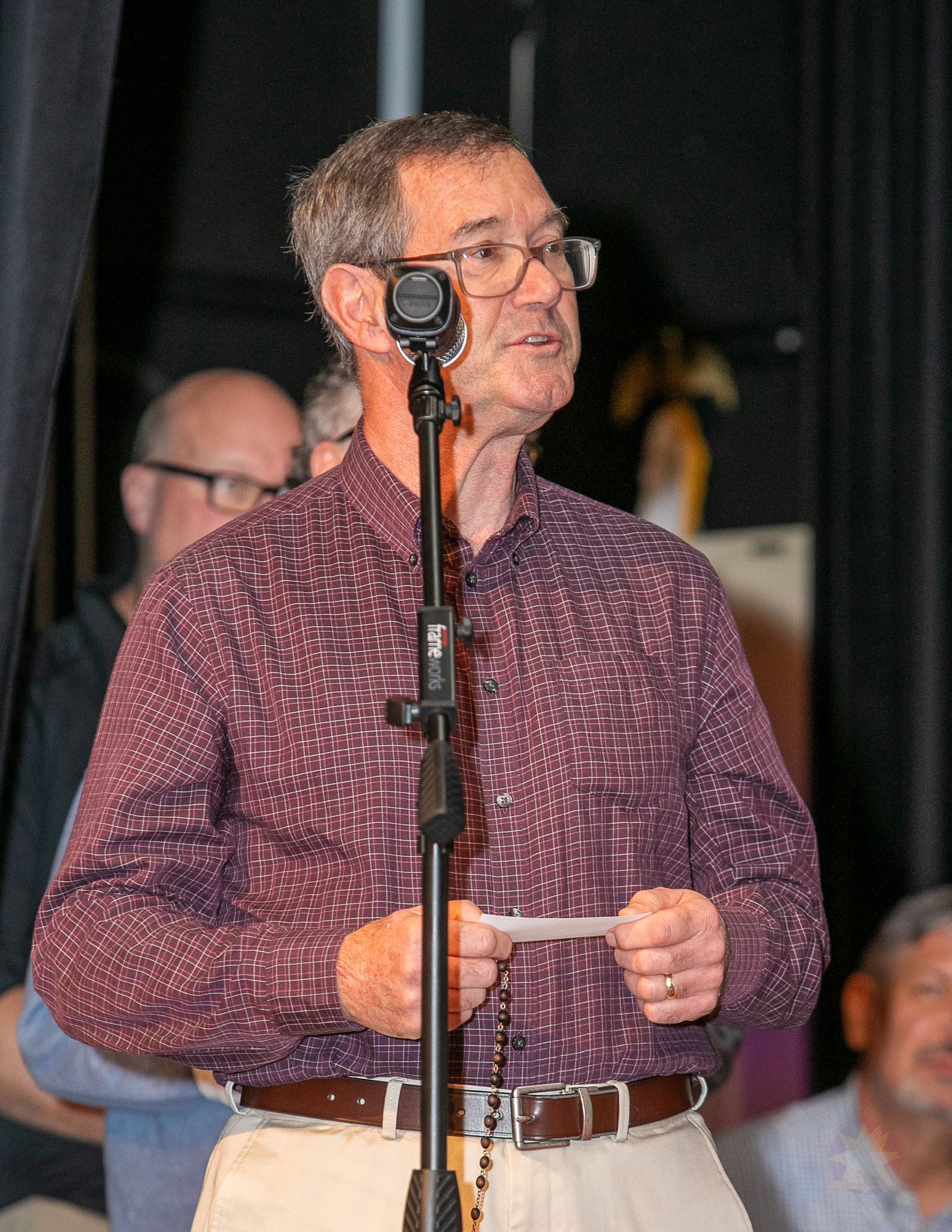 Man speaking at a microphone onstage, wearing a maroon patterned shirt and khaki pants.