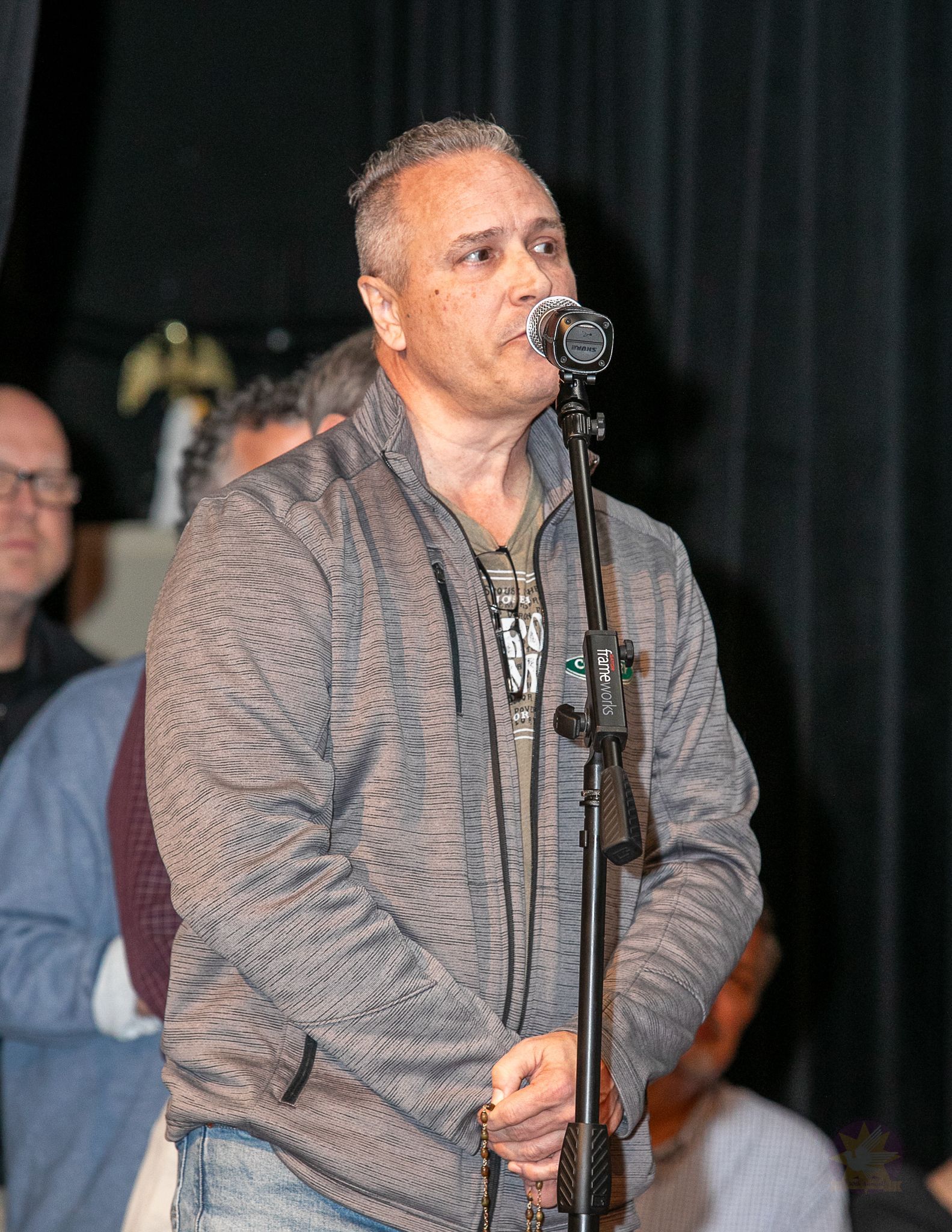 Man speaking into a microphone on stage, wearing a patterned sweater with musicians blurred in the background