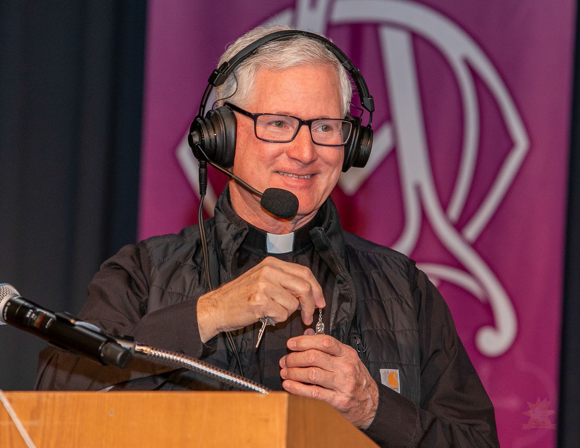 Man speaking at a podium with a headset microphone, holding a small object, against a purple backdrop