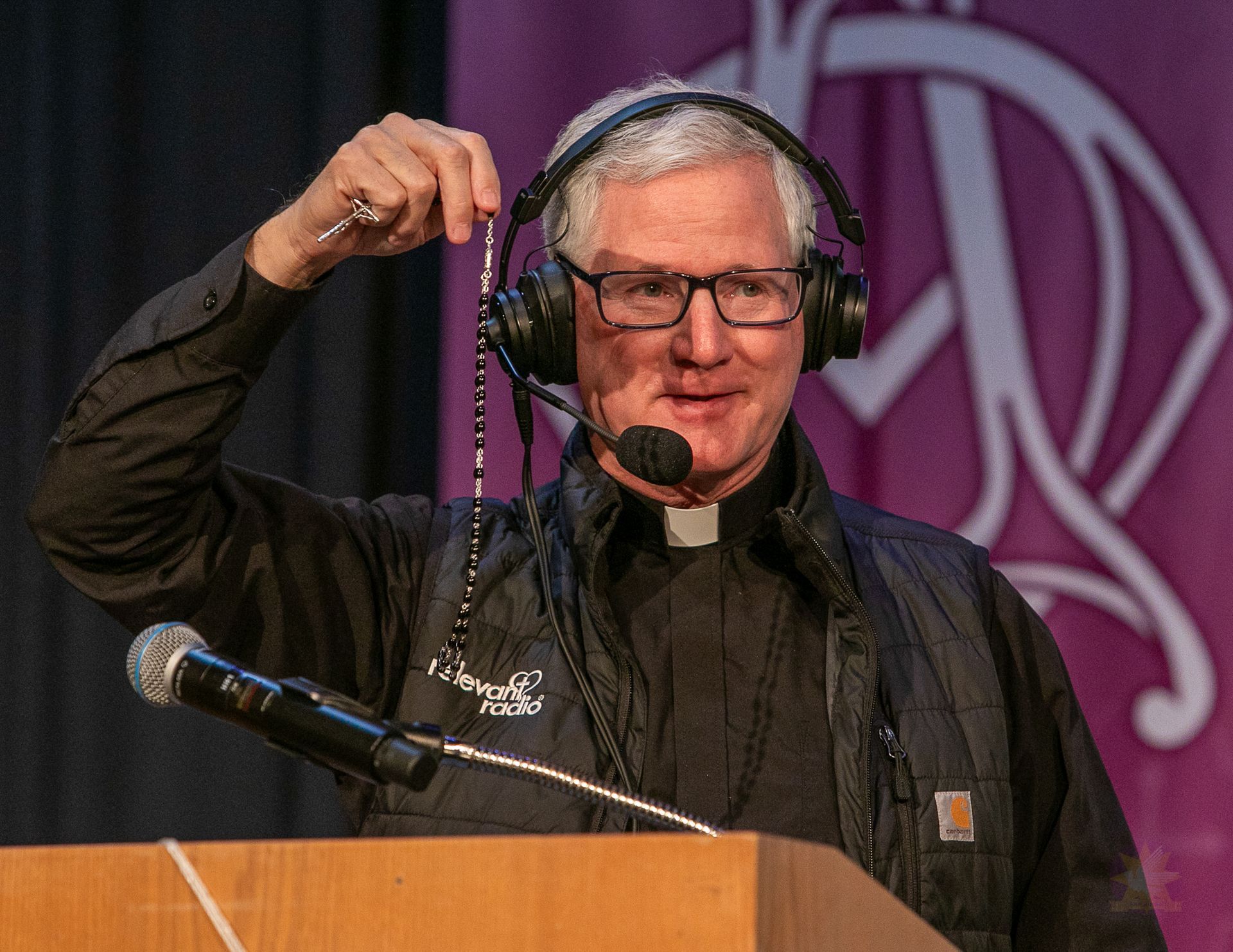 Fr. Rocky adjusting headphones at a podium with microphones on a purple-lit stage