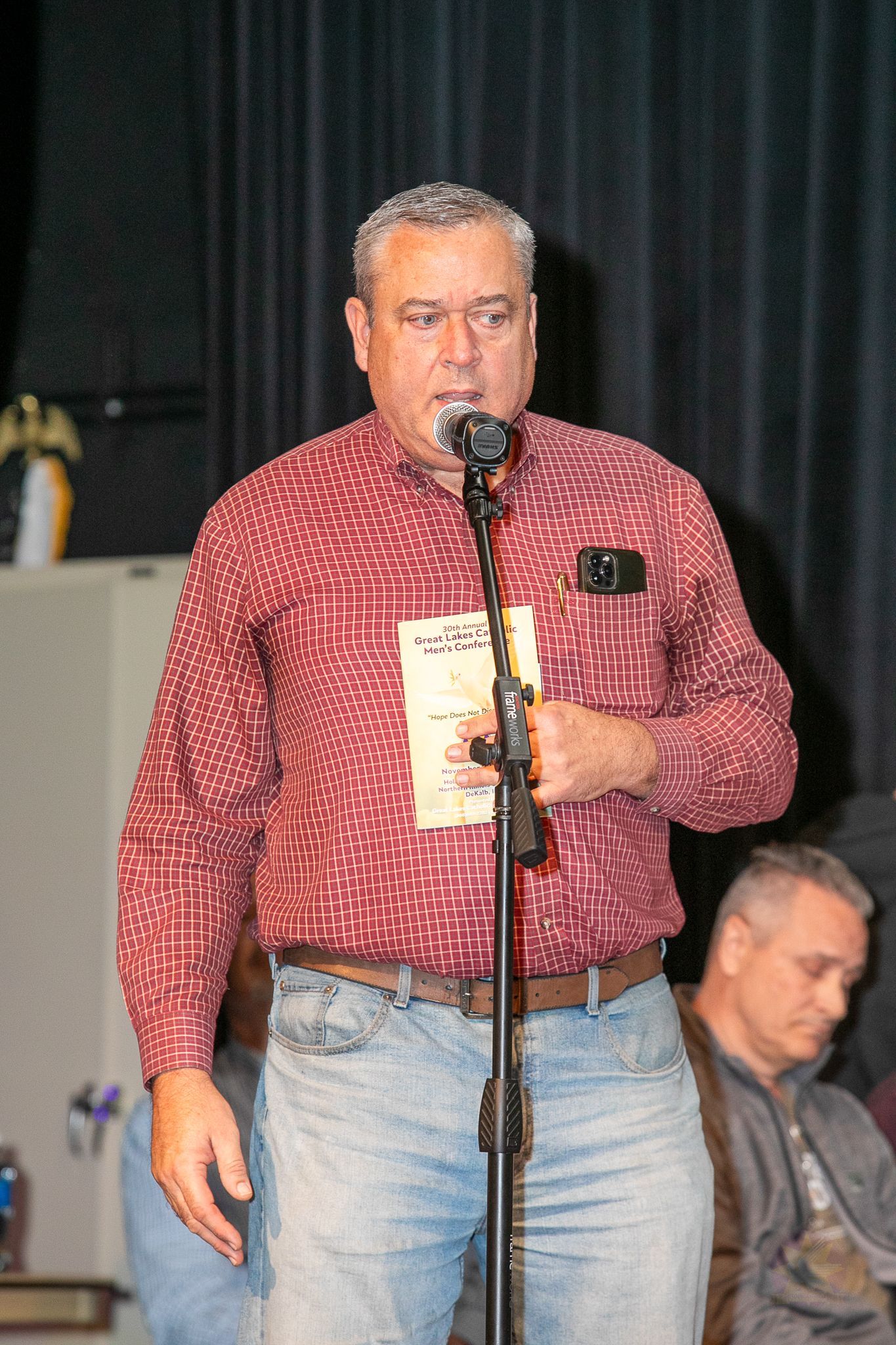 Man speaking at a microphone onstage, wearing a red patterned shirt and jeans, holding a cup.