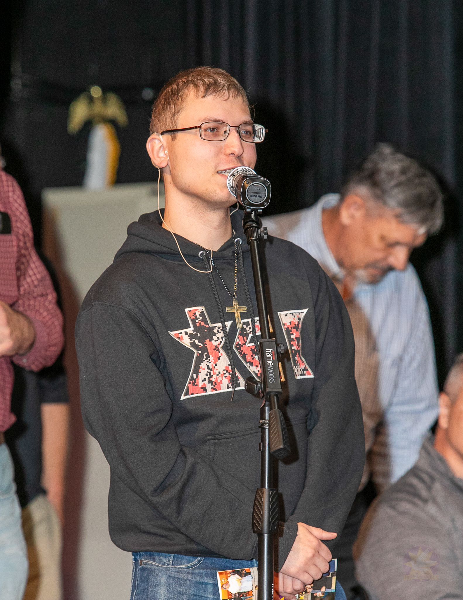 Person speaking at a microphone on a stage, wearing a dark hoodie and glasses, with others in the background.