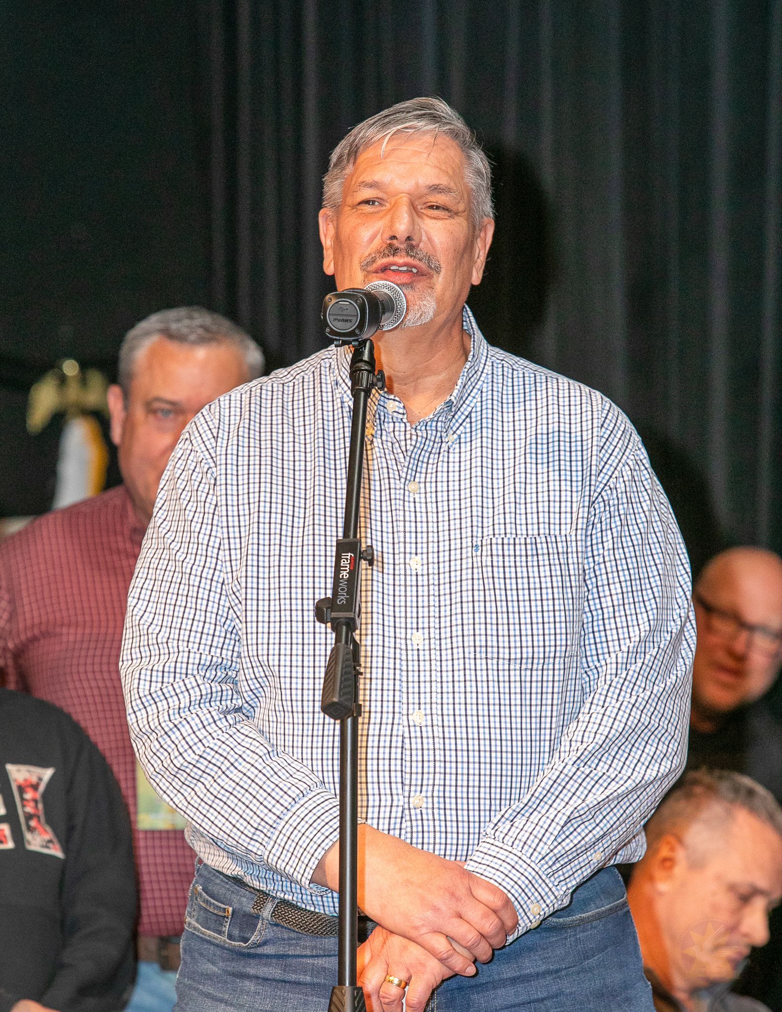 Man speaking at a microphone on stage, wearing a blue plaid shirt, with several people in the background.