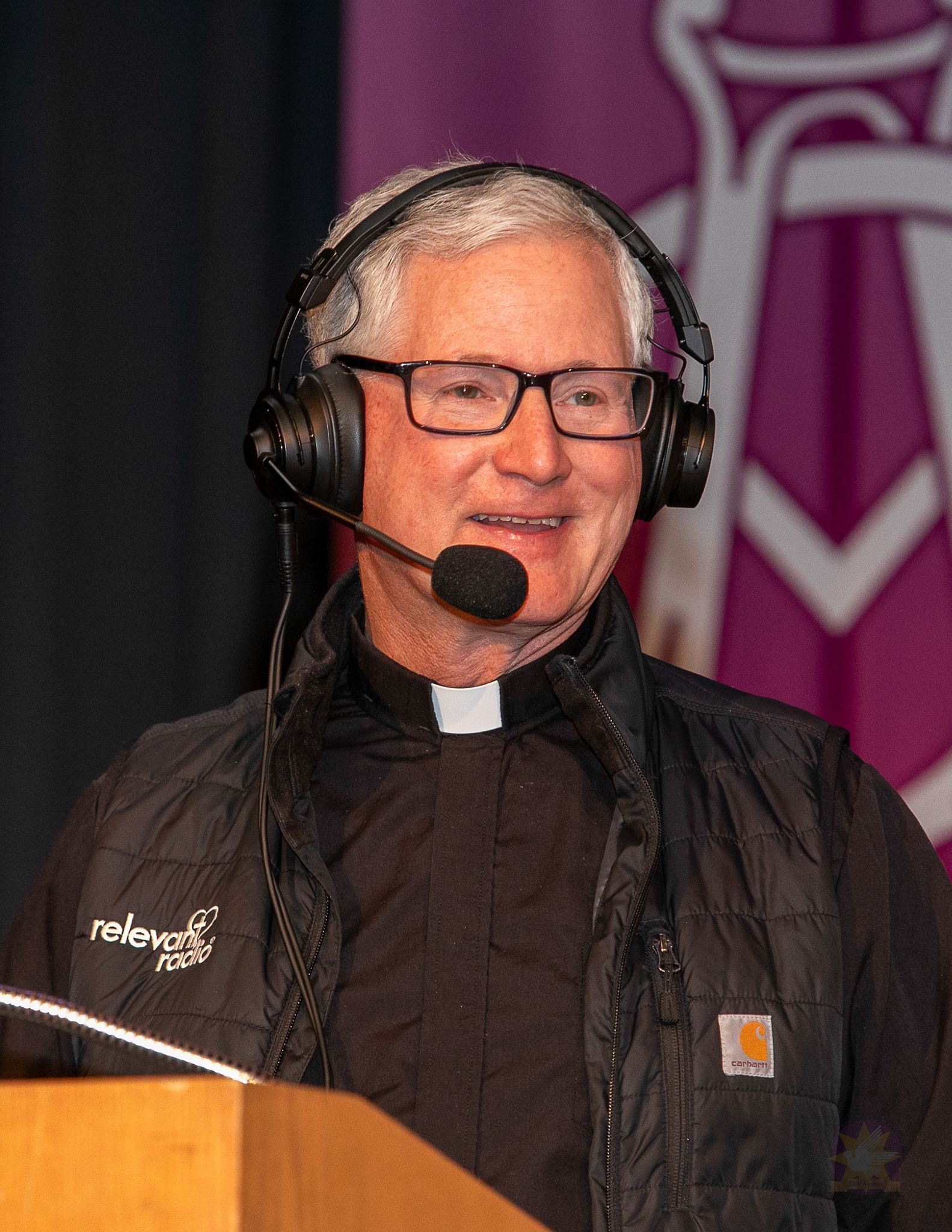 Man in clerical attire wearing a headset microphone, speaking at a podium with a purple banner behind him