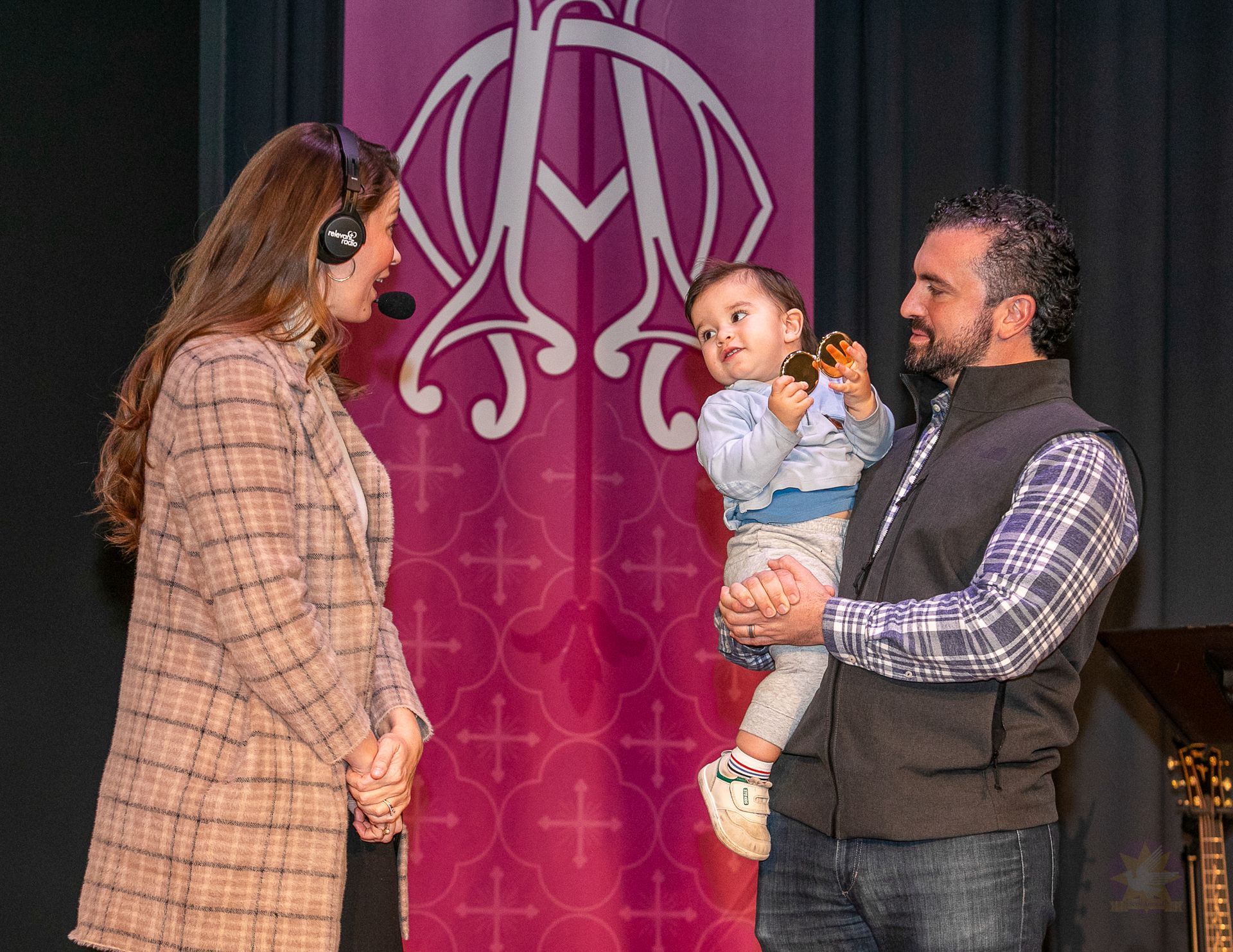 Three people on stage; a man holds a baby while a woman faces them near a pink backdrop with a monogram.