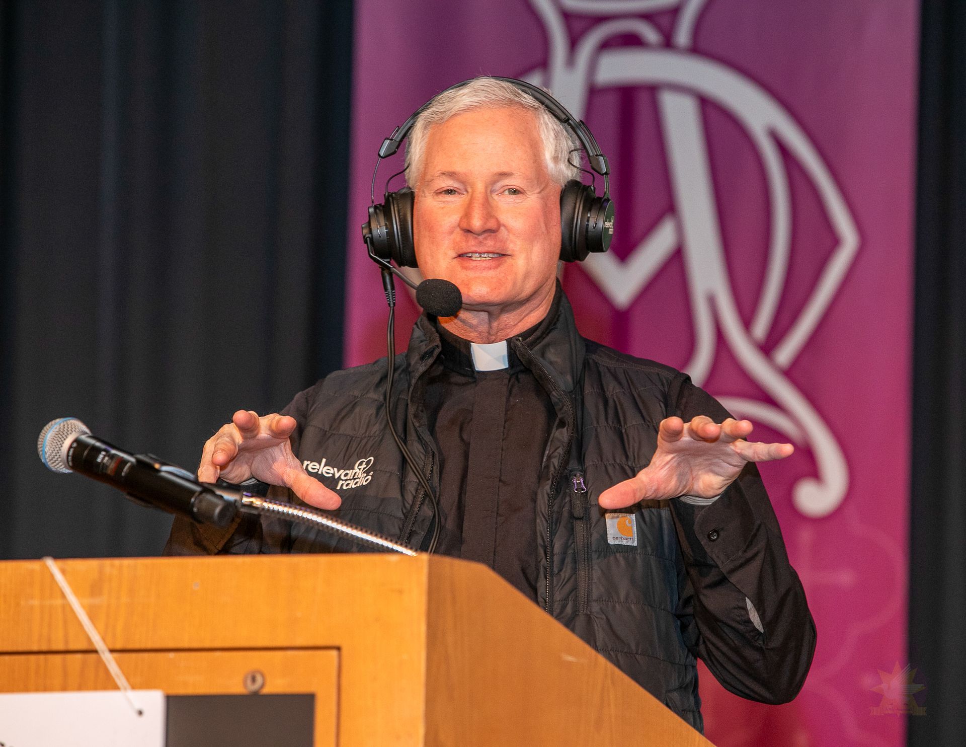 Fr. Rocky speaking at a podium with a headset microphone, hands raised, in front of a pink backdrop
