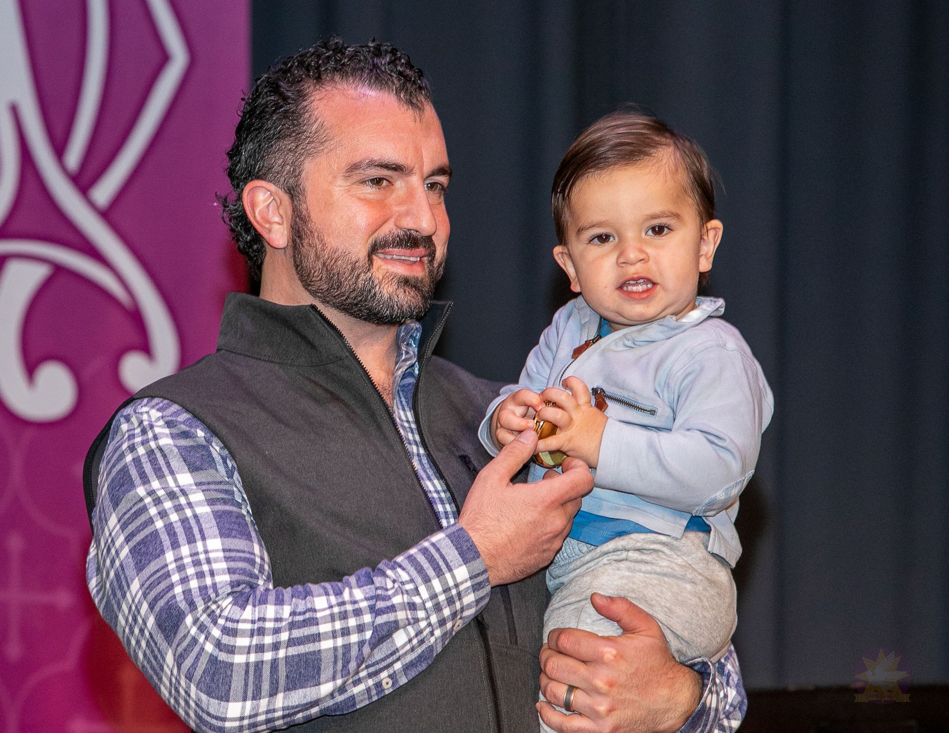 Man holding a toddler on stage, both facing the camera, with a dark backdrop and purple banner.