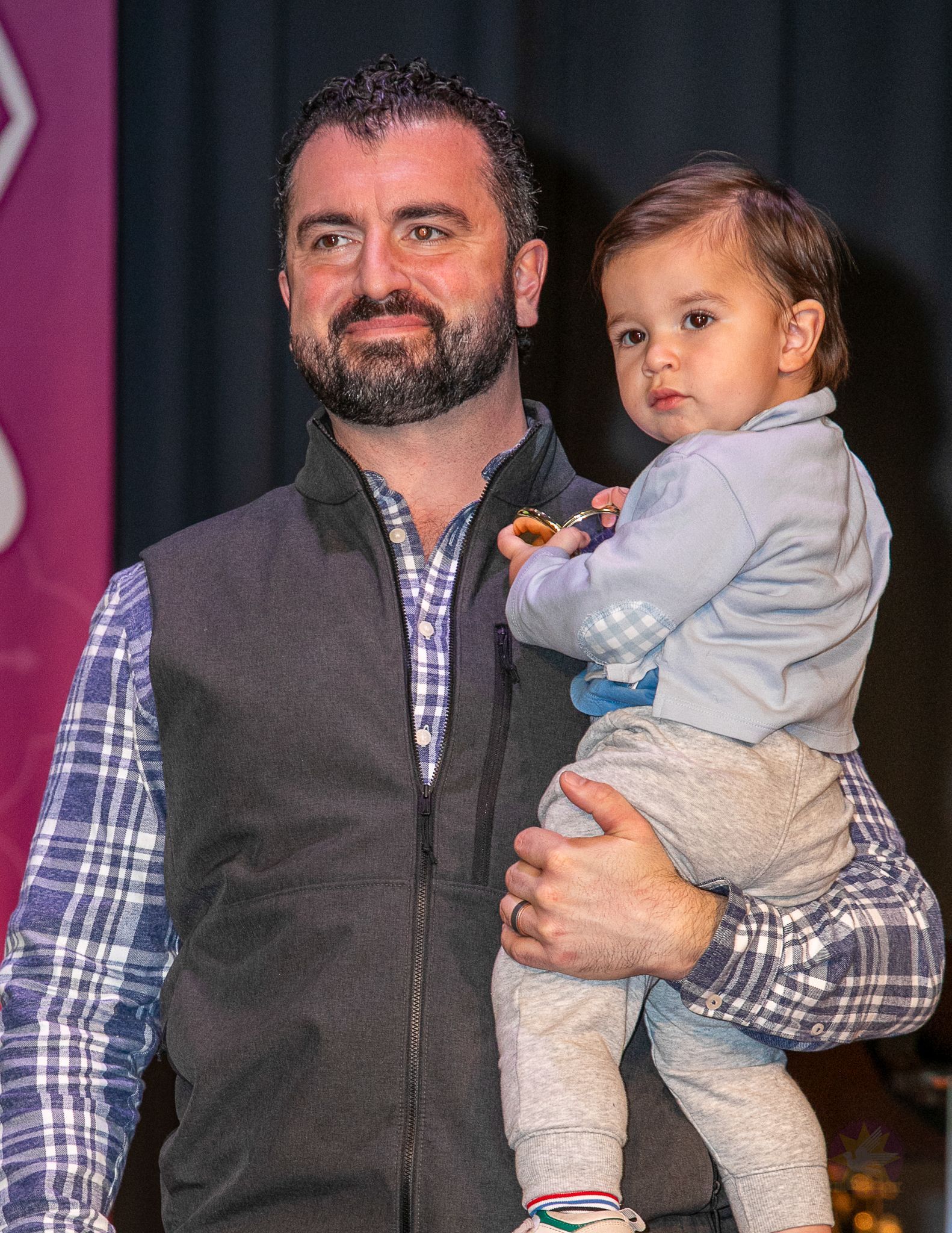 Man holding a toddler onstage, both facing the camera, with dark curtains in the background.