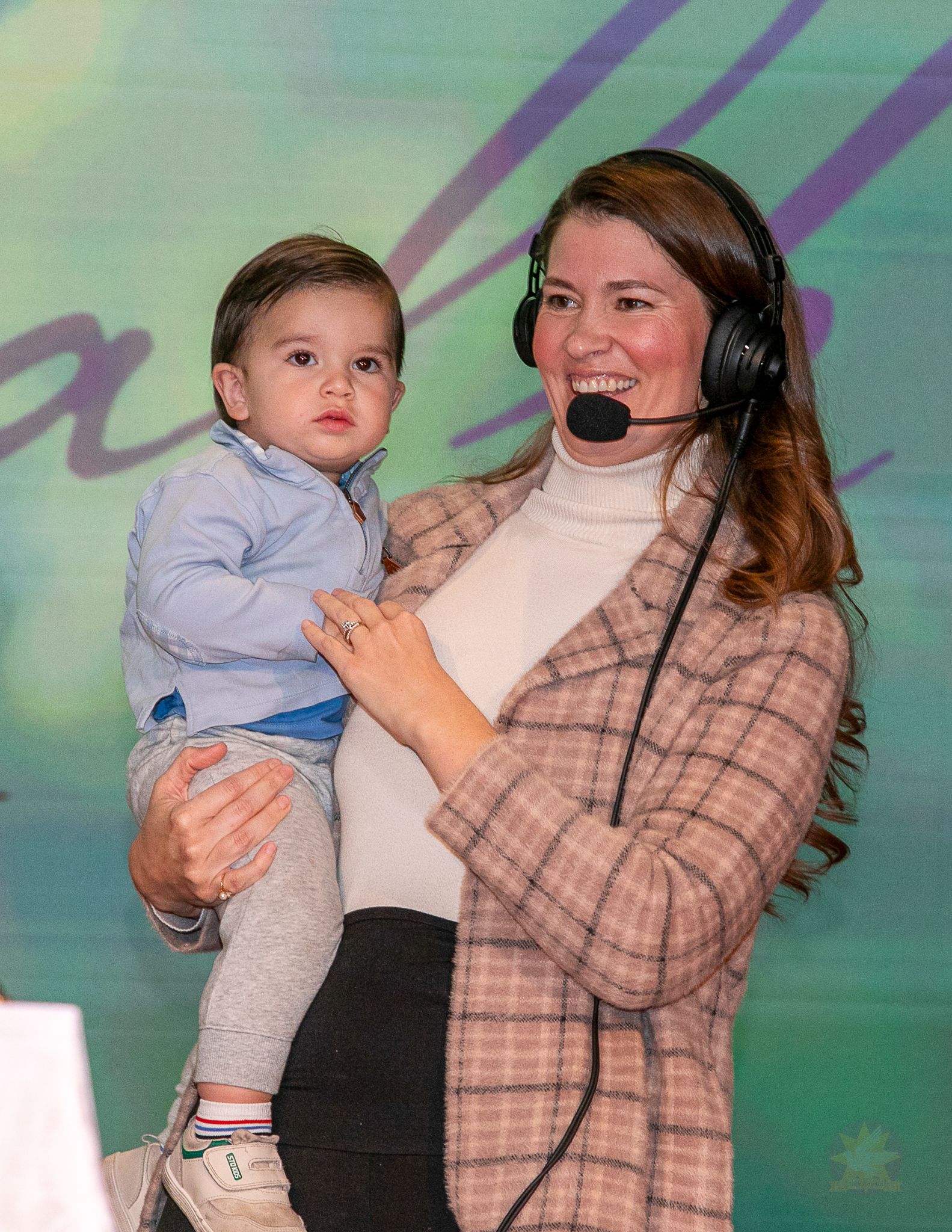 Woman wearing a headset holds a child against a green backdrop.