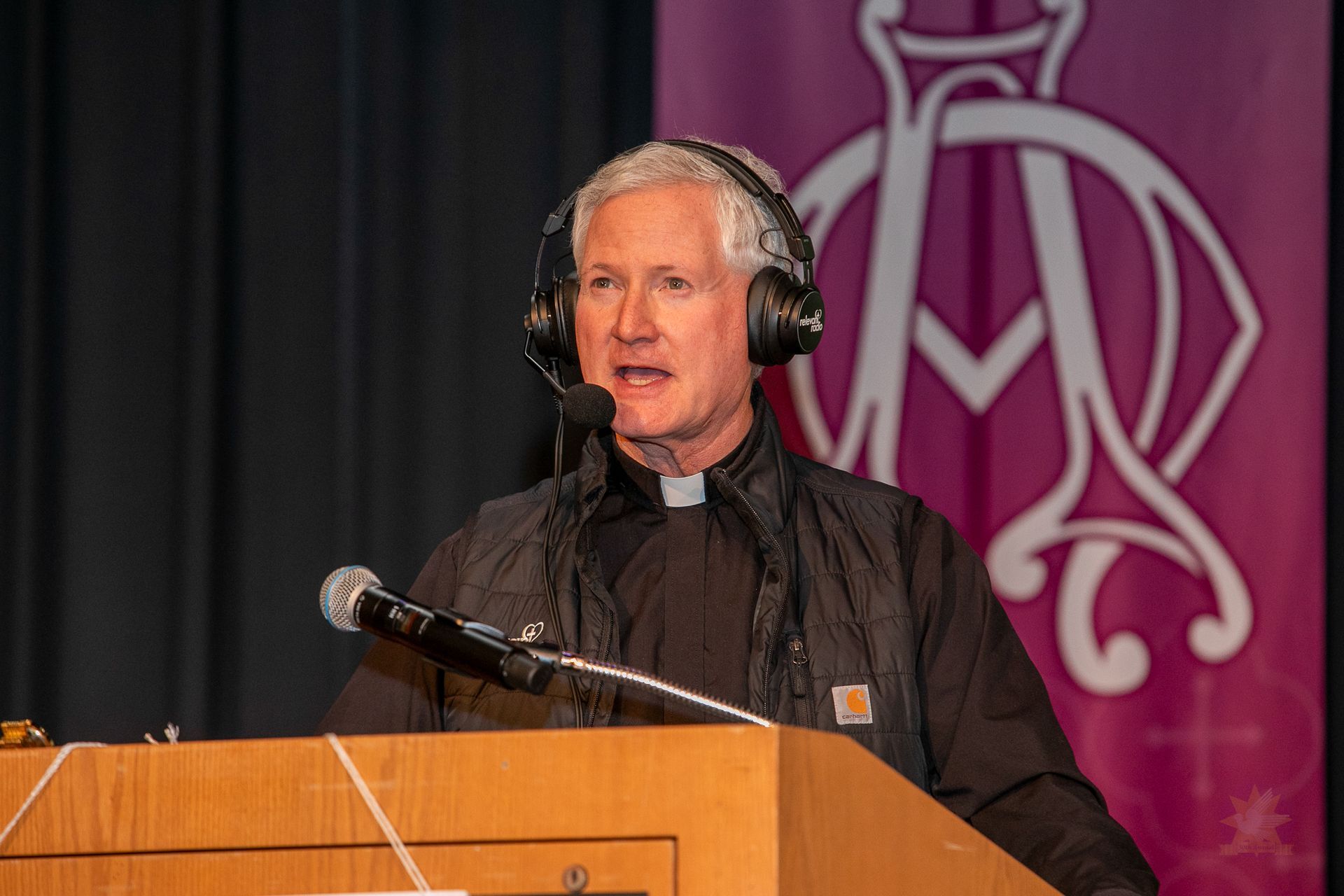 Speaker in black jacket and headset at a podium, with a microphone and purple backdrop behind him.