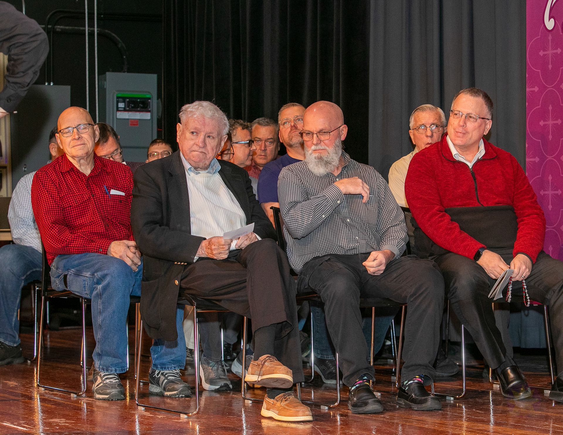 Five men seated on chairs on a stage, with one speaking into a microphone and others listening.