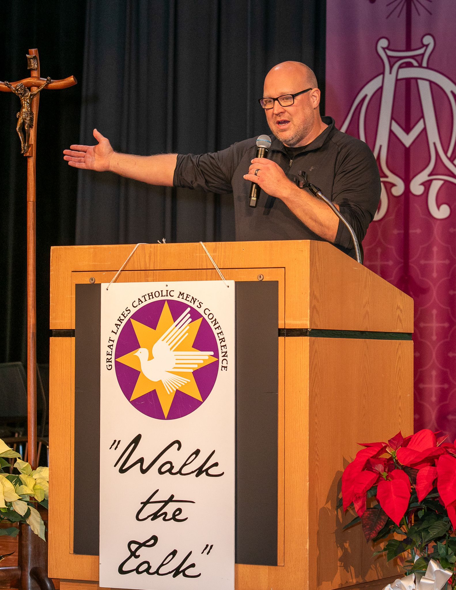 Man speaking at a church podium with a “Walk the Talk” sign, gesturing beside a cross and purple banner