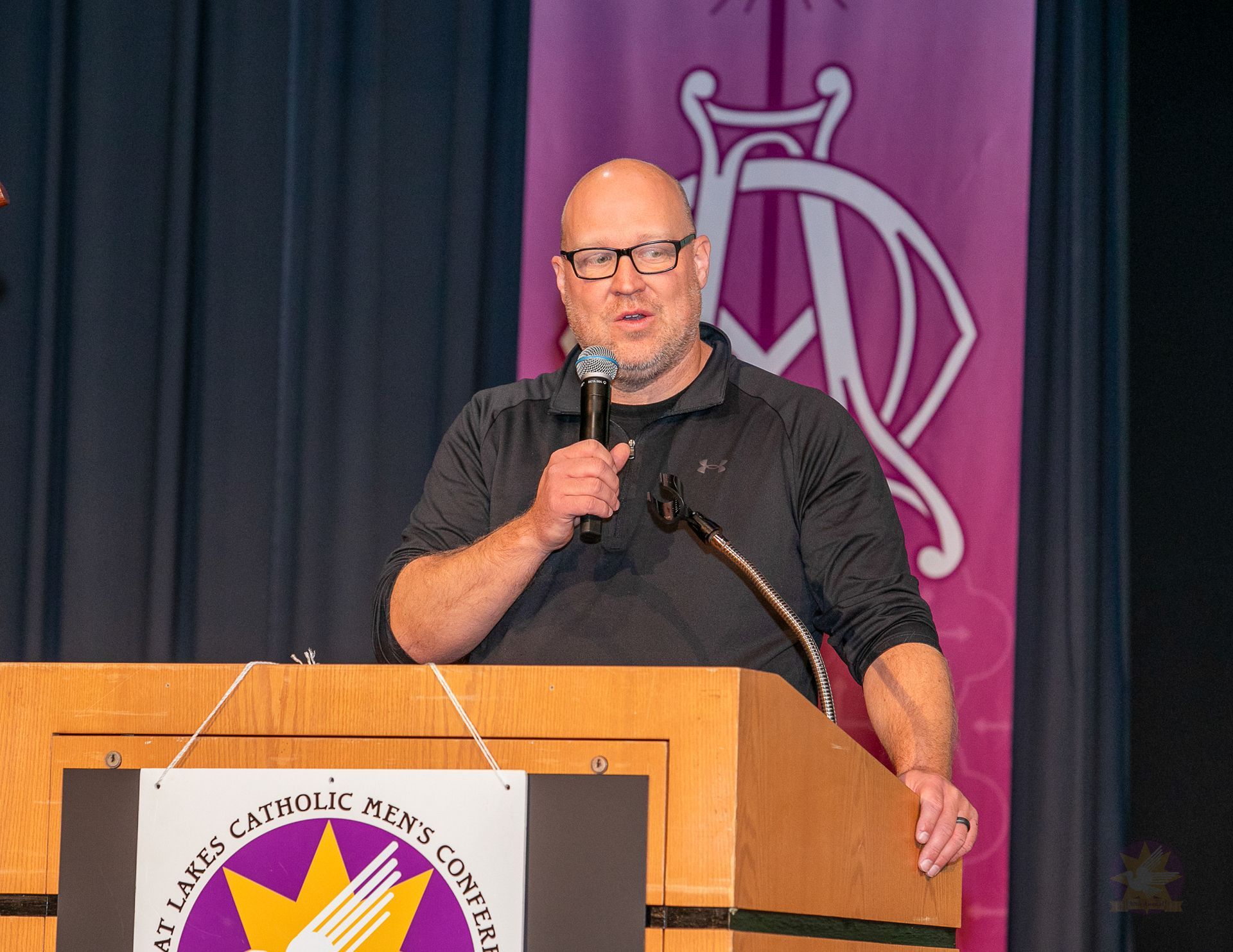 Mitch Striedl at podium holding microphone on stage with dark curtains and purple banner backdrop.