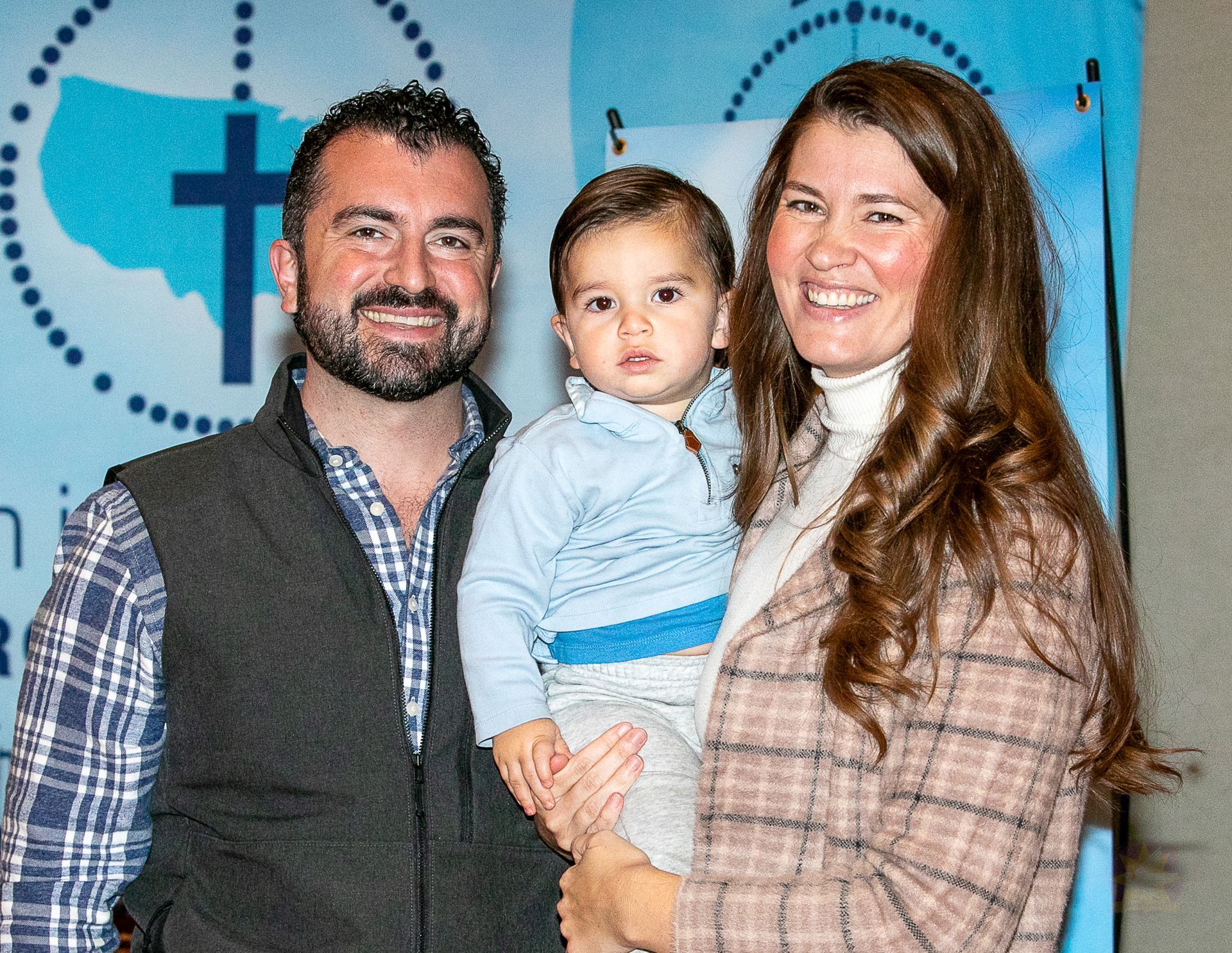 Three people posing together in front of a blue event backdrop, smiling at the camera.