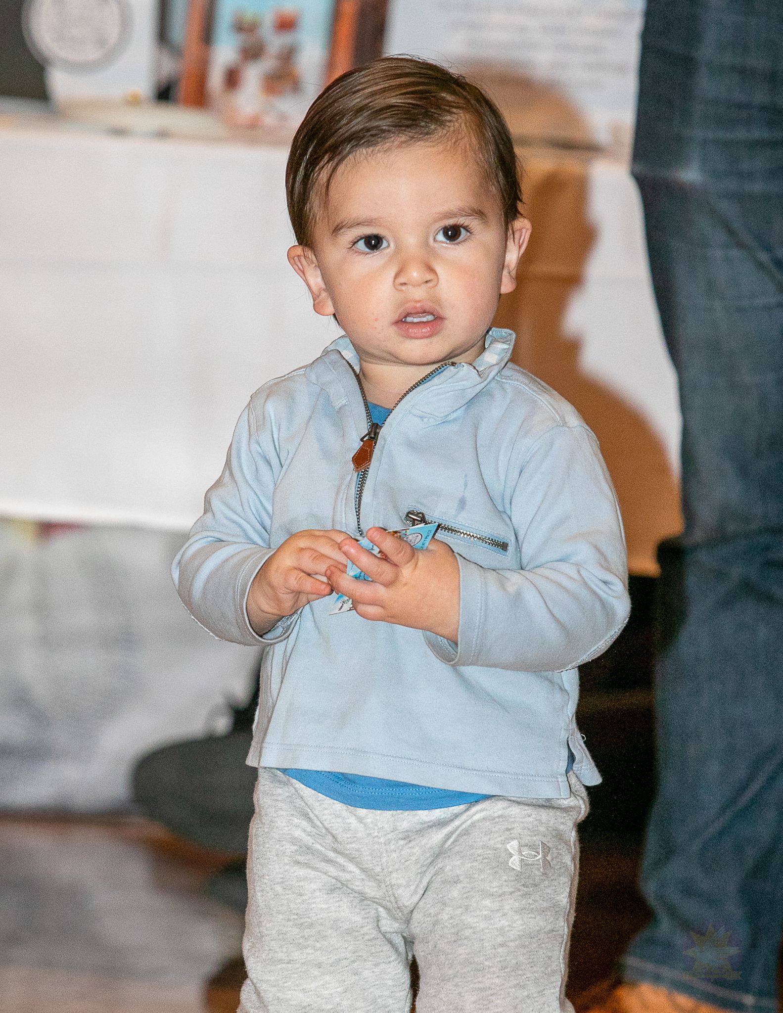 Child standing indoors in a light blue sweater and gray pants, looking at the camera.