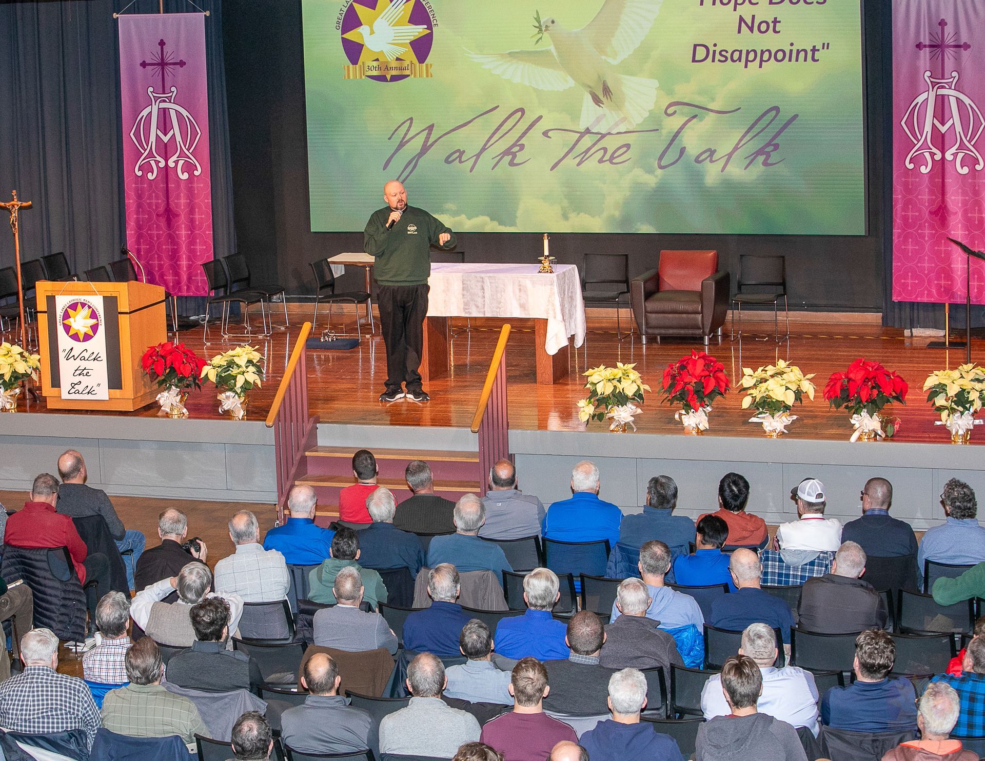 Speaker on church stage addressing seated audience with floral decorations and large banner backdrop
