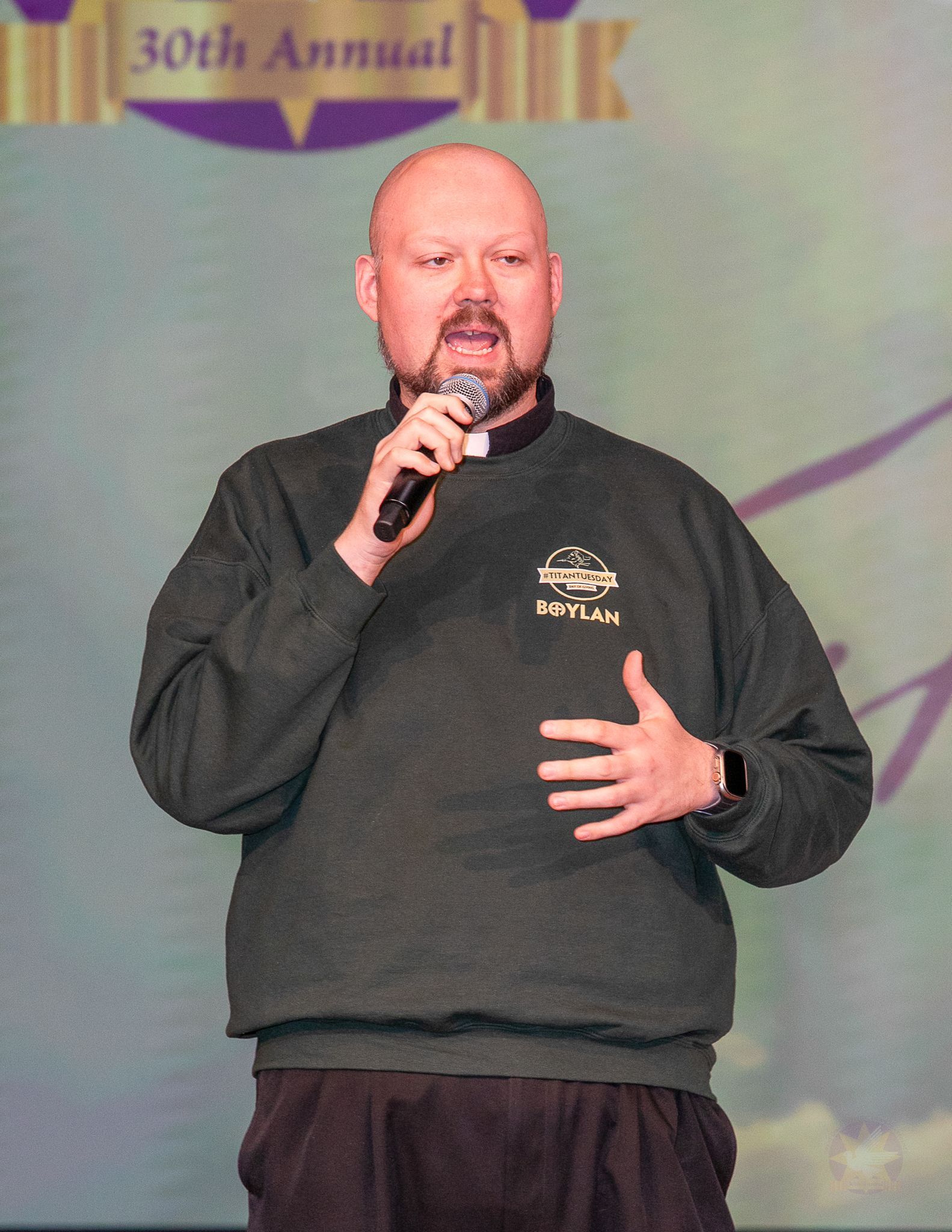 Rev. Robert Blood speaking into a microphone on stage, wearing a black sweatshirt against a gray backdrop.